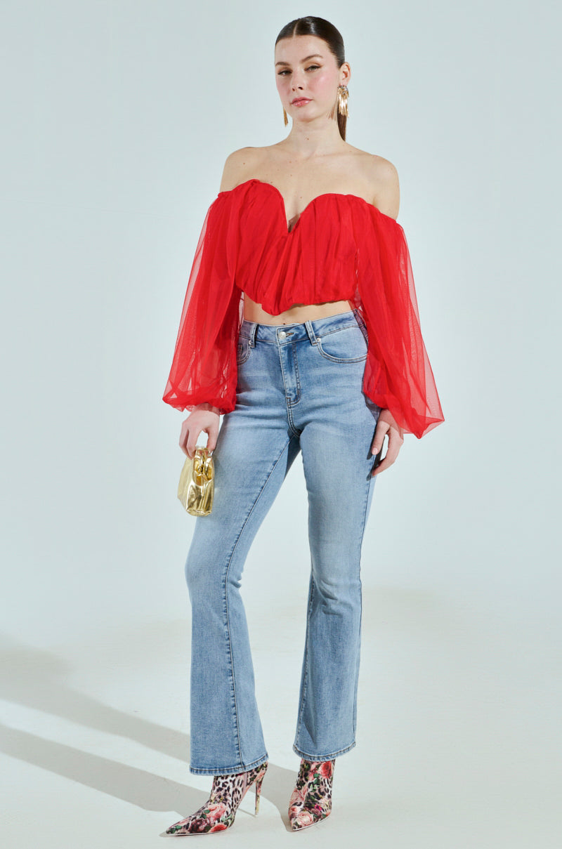 A person stands against a light background wearing the "FLY WITH ME CORSET TOP," a red off-the-shoulder design with sheer long sleeves. They pair it with blue jeans, leopard print shoes, hold a small gold bag, and wear long earrings.