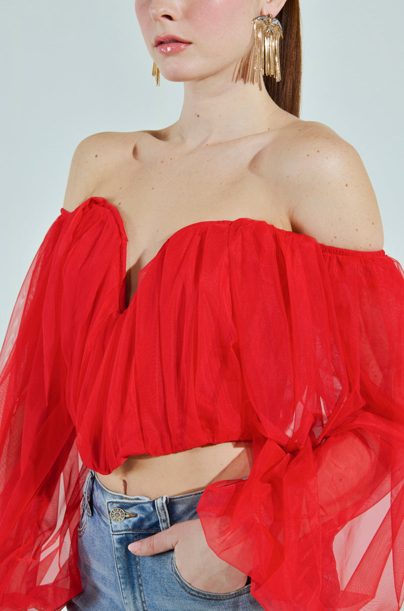 A woman in the sheer, red "FLY WITH ME CORSET TOP" with puffy sleeves and a sweetheart neckline is against a neutral background. Large gold earrings and denim jeans complement the look, while her face remains partially obscured.