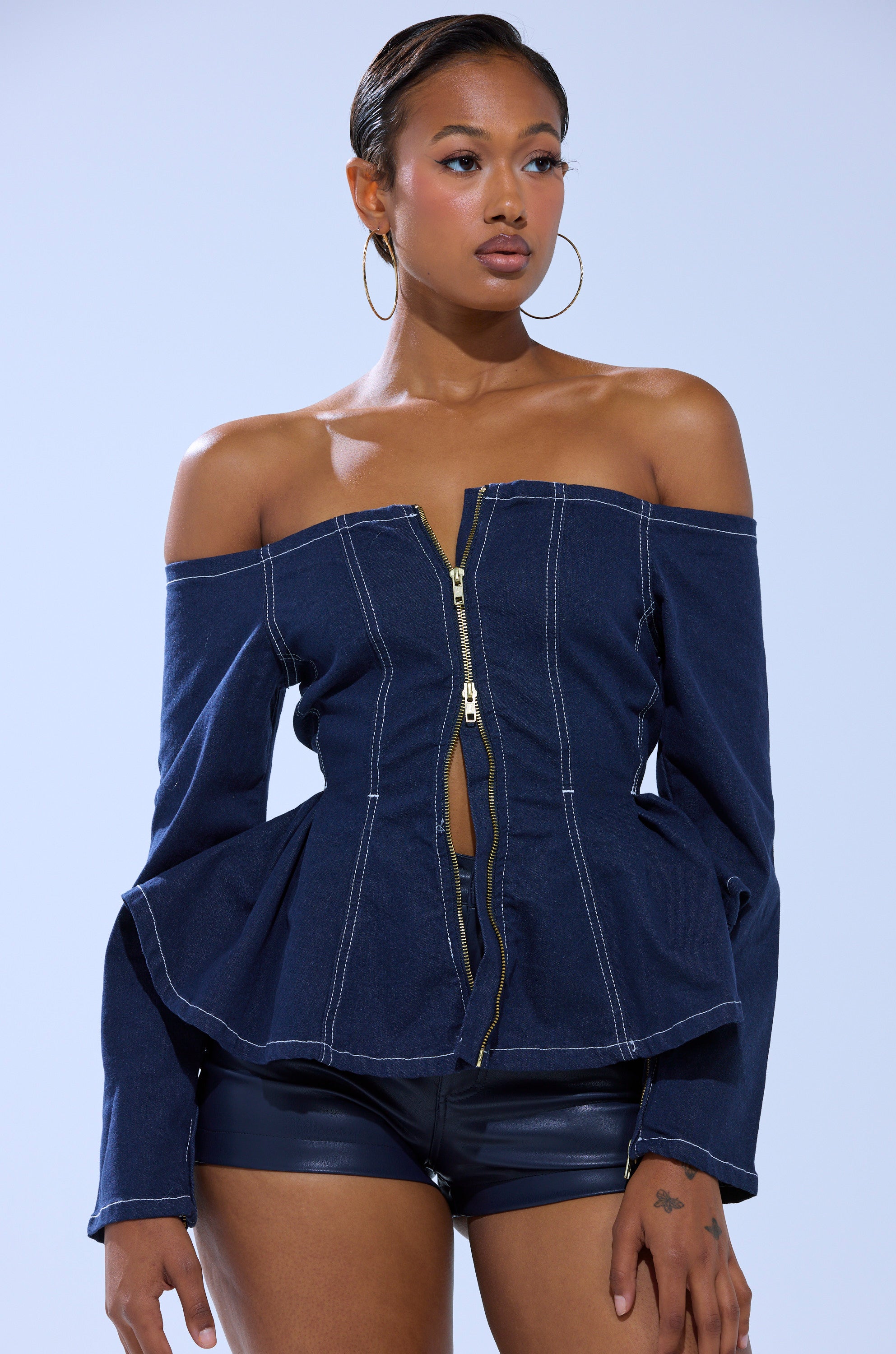 A woman with her hair pulled back and a tattoo on her left hand wears large hoop earrings and the DENIM FOR DAYS ZIP UP BLOUSE, paired with black leather shorts, as she stands against a light blue background.