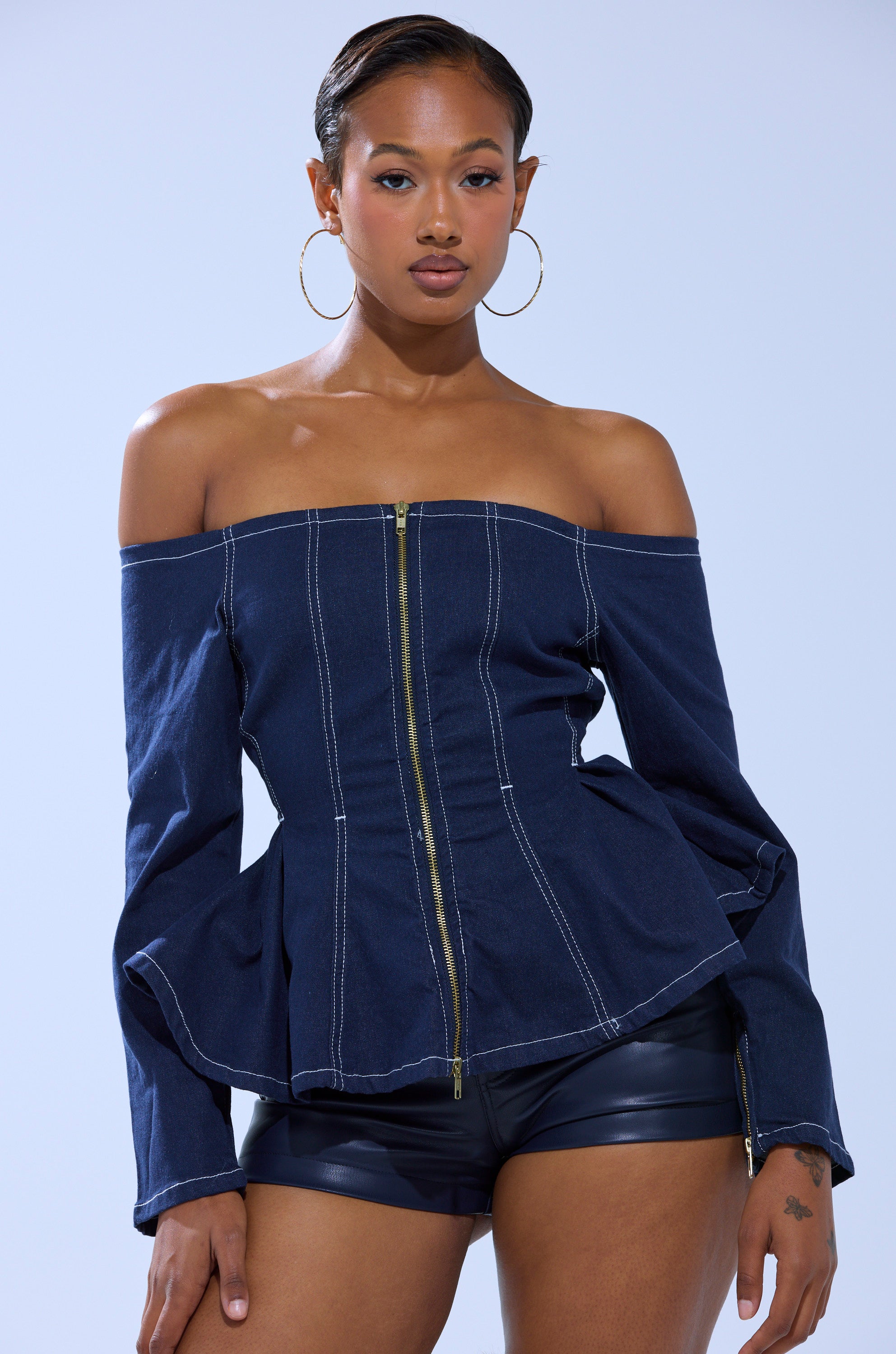A woman poses confidently against a plain light blue background, wearing large hoop earrings, black leather shorts, and the DENIM FOR DAYS ZIP UP BLOUSE, a strapless denim top with white stitching.