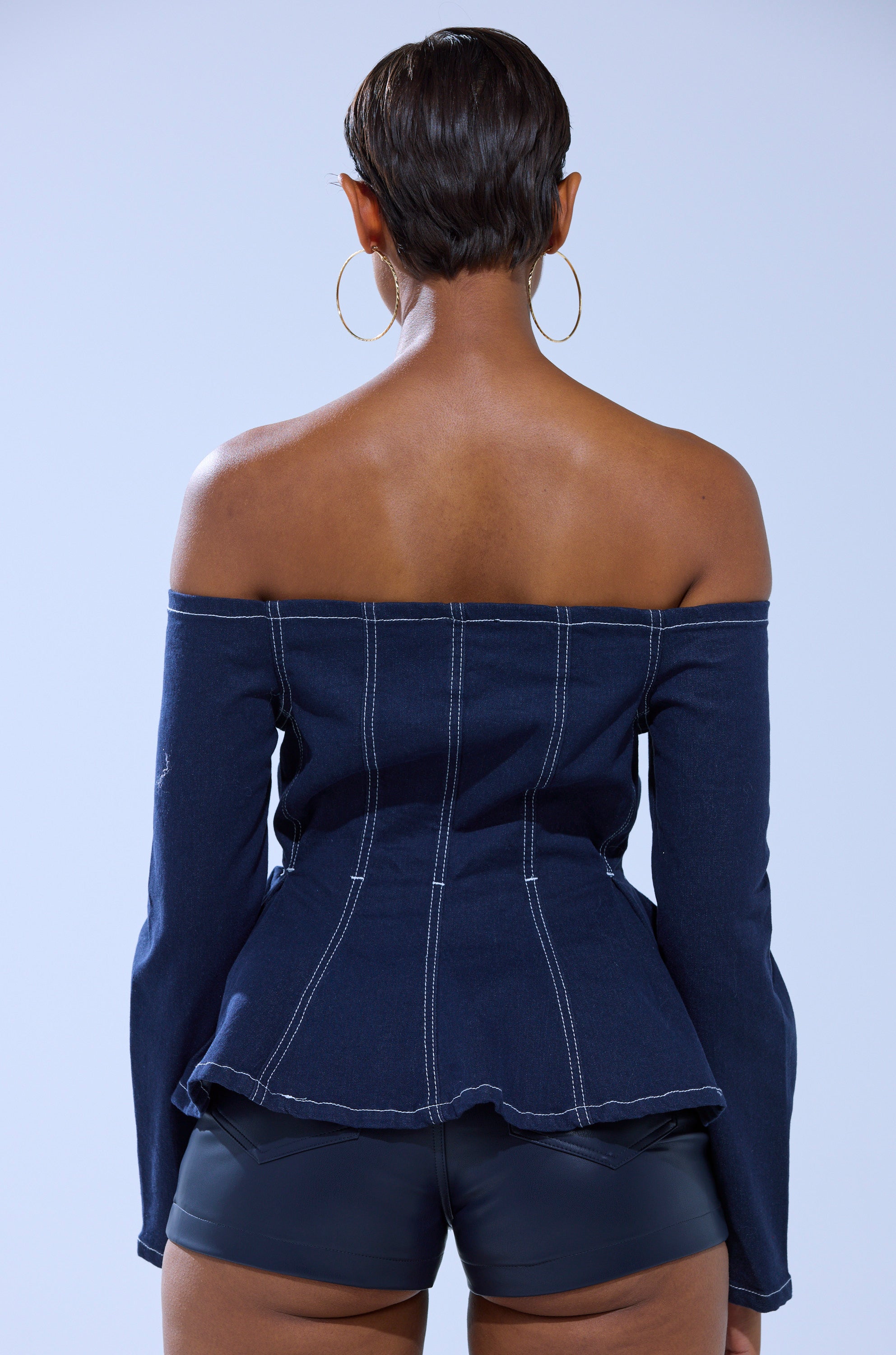 A person with short hair, wearing large hoop earrings and black shorts, is seen from the back against a plain light background, wearing the DENIM FOR DAYS ZIP UP BLOUSE featuring dark wash denim and visible white stitching.