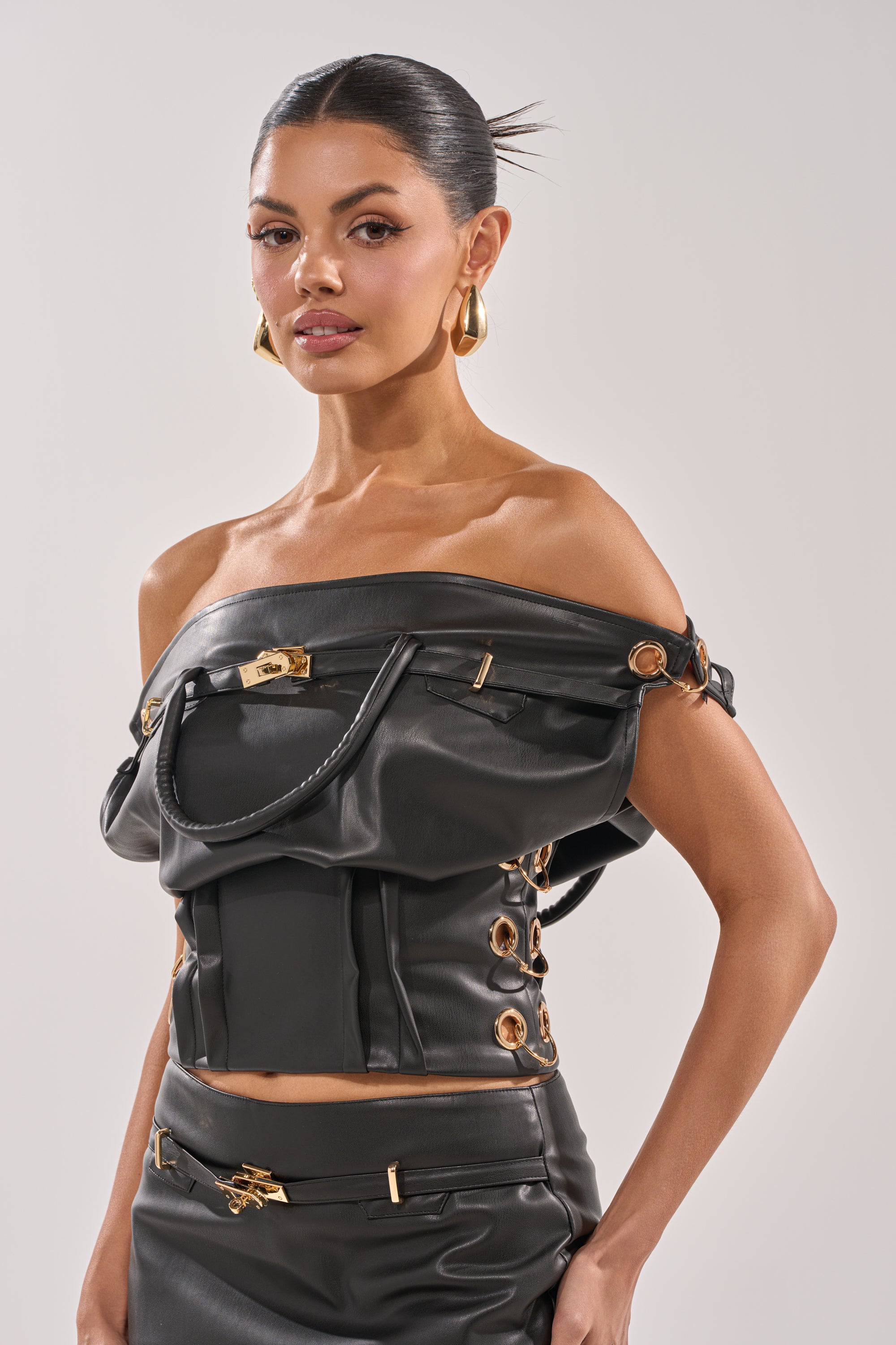 A woman with slicked-back hair wears the MONEY TALKS OFF THE SHOULDER TOP in black faux leather with gold-tone hardware and a matching skirt, accessorized with large gold hoop earrings, against a plain light background. 2