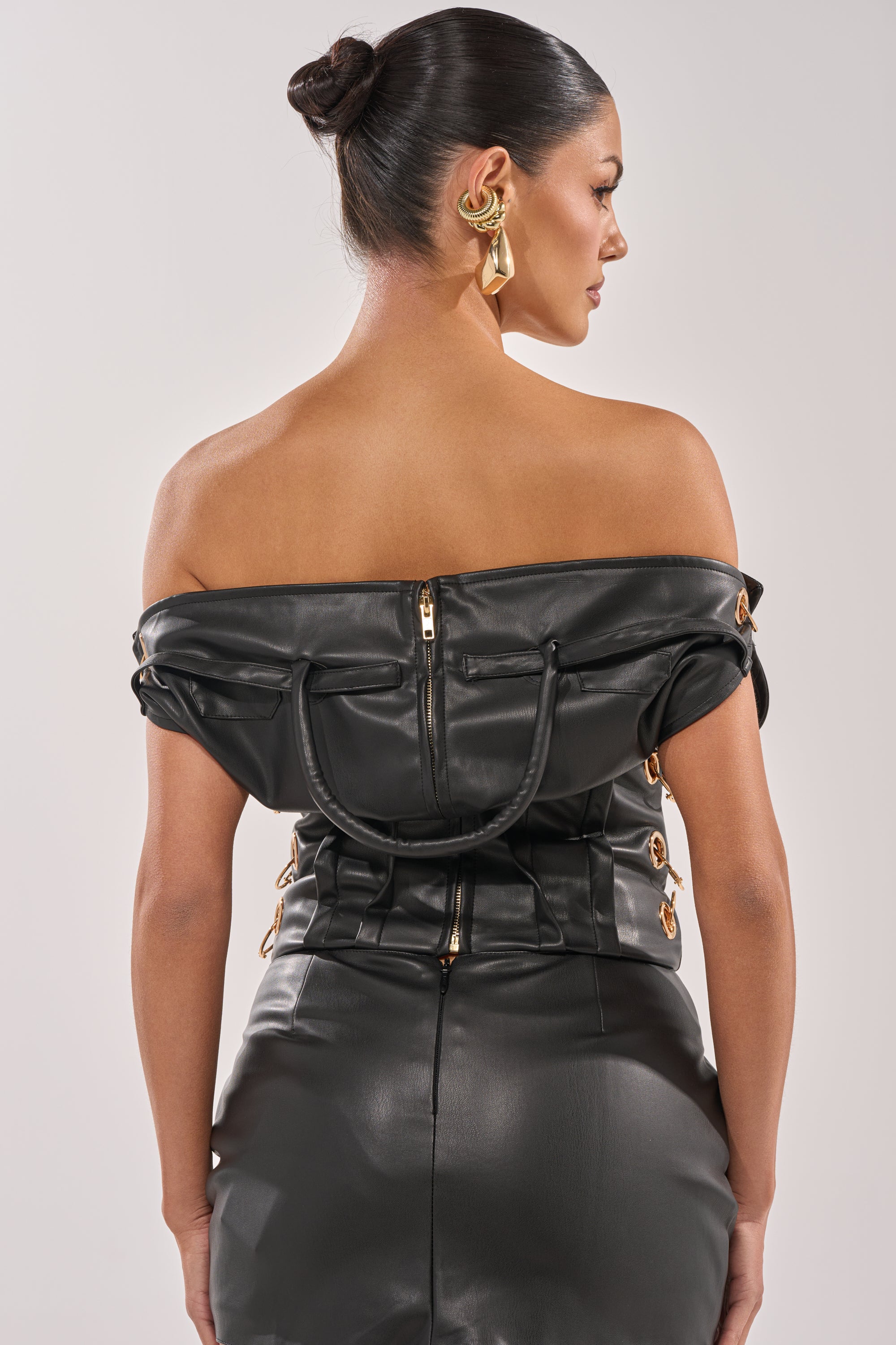 A woman with dark hair in a bun wears the MONEY TALKS OFF THE SHOULDER TOP, featuring a black purse-inspired silhouette with gold-tone hardware and back zipper detail. She stands slightly turned, showcasing her back. 7