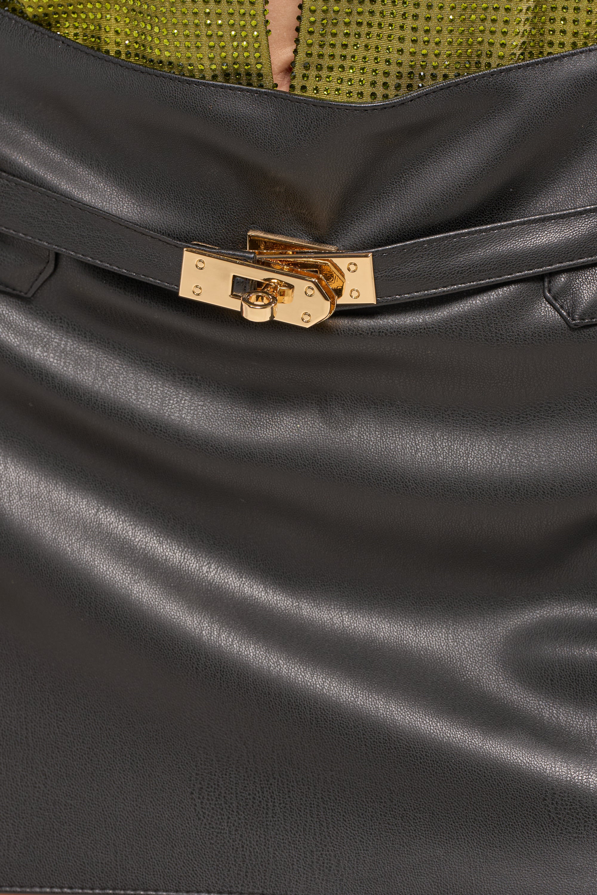A close-up of the MONEY TALKS MINI SKIRT in black faux leather beside a black leather handbag with a gold clasp, all set against green fabric with shiny embellishments. 6
