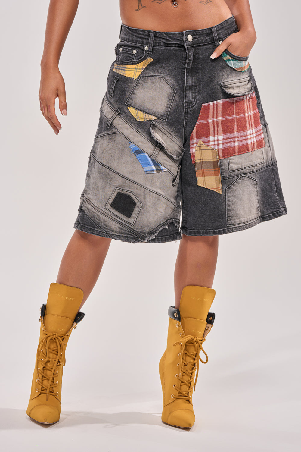 A person wears DREA MIXED PLAID BERMUDA SHORTS and yellow lace-up ankle boots. The knee-length shorts feature colorful patchwork. One hand is in a pocket. Only the torso and legs are shown against a plain light background. 1
