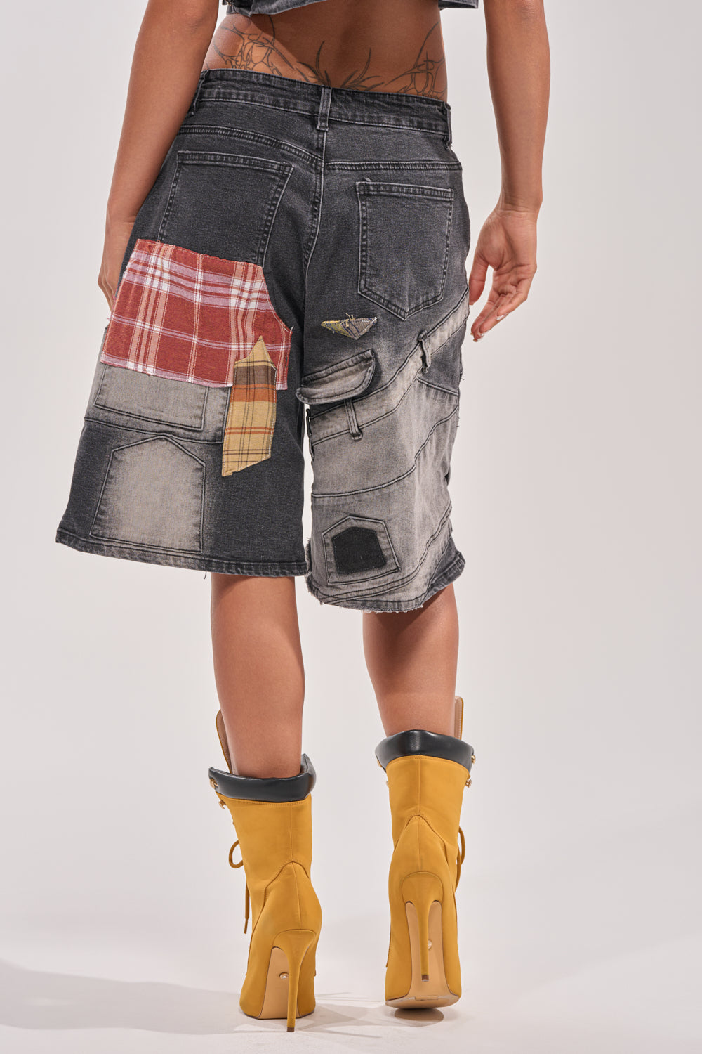 A person faces away, showcasing the DREA MIXED PLAID BERMUDA SHORT—black denim with red plaid accents—paired with mustard-yellow heeled boots. Tattoos on their lower back and arms stand out against a plain light background. 5