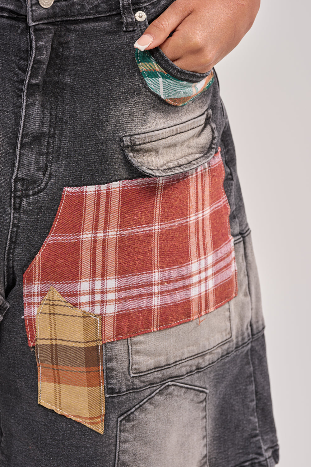 A close-up of someone wearing the DREA MIXED PLAID BERMUDA SHORT, featuring black denim with faded areas, bold red and yellow plaid patches, and a green plaid-lined pocket for a standout street style look. 8