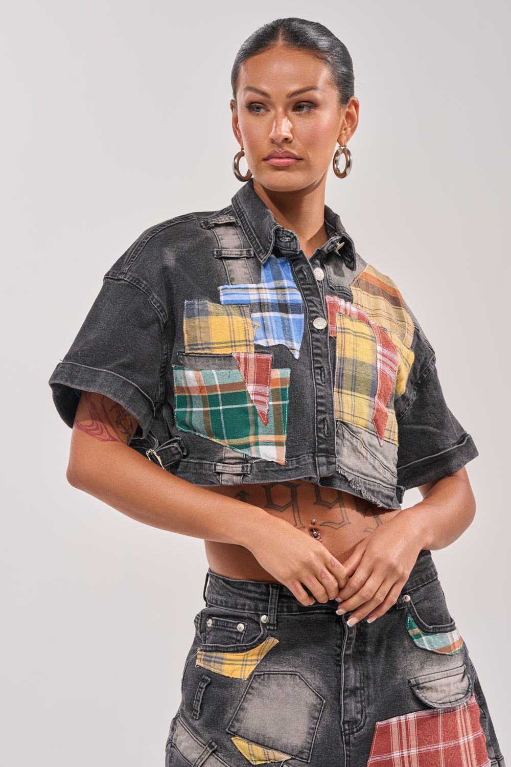 Wearing the DREA MIXED PLAID CROP SHORT SLEEVE BUTTON DOWN, a woman channels street style with hoop earrings, slicked-back hair, and a confident look against a plain background. 0