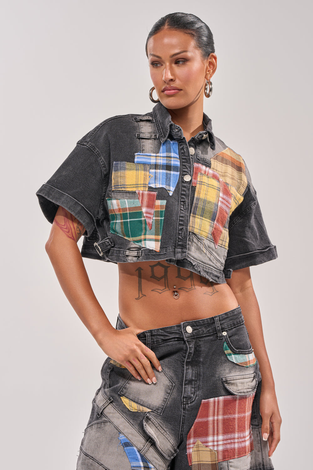 A woman stands confidently with one hand on her hip, showing torso tattoos while wearing the DREA MIXED PLAID CROP SHORT SLEEVE BUTTON DOWN and matching jeans with colorful patchwork designs. 1