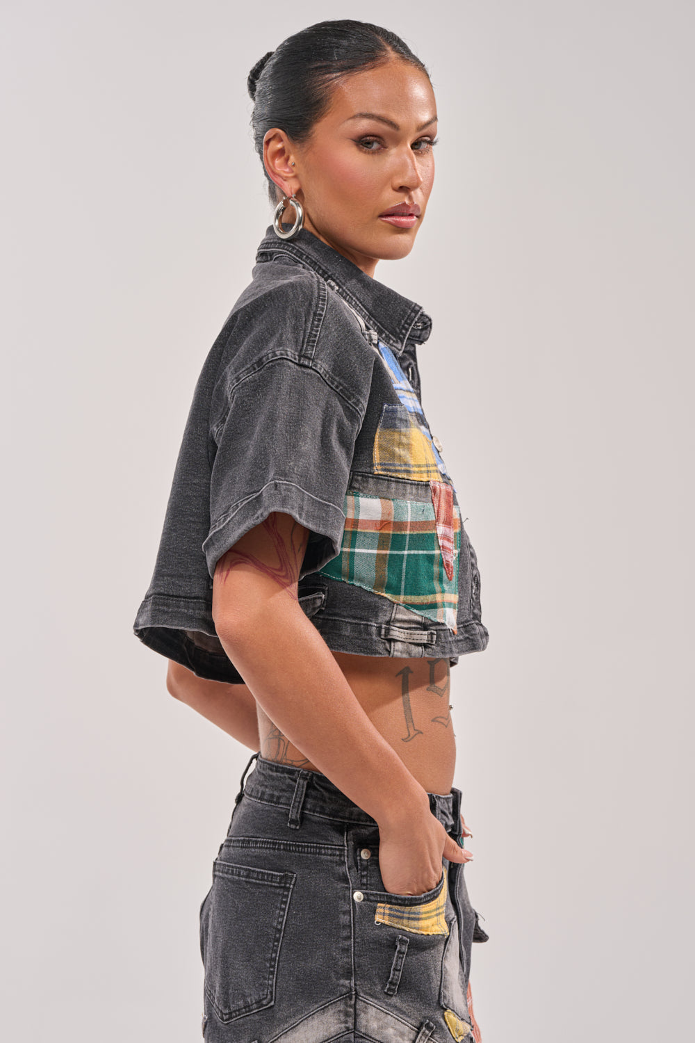 A woman with long dark hair in a bun, hoop earrings, and tattoos stands confidently in the DREA MIXED PLAID CROP SHORT SLEEVE BUTTON DOWN—a black denim crop shirt with plaid patchwork—paired with black jeans and one hand in her pocket. 2