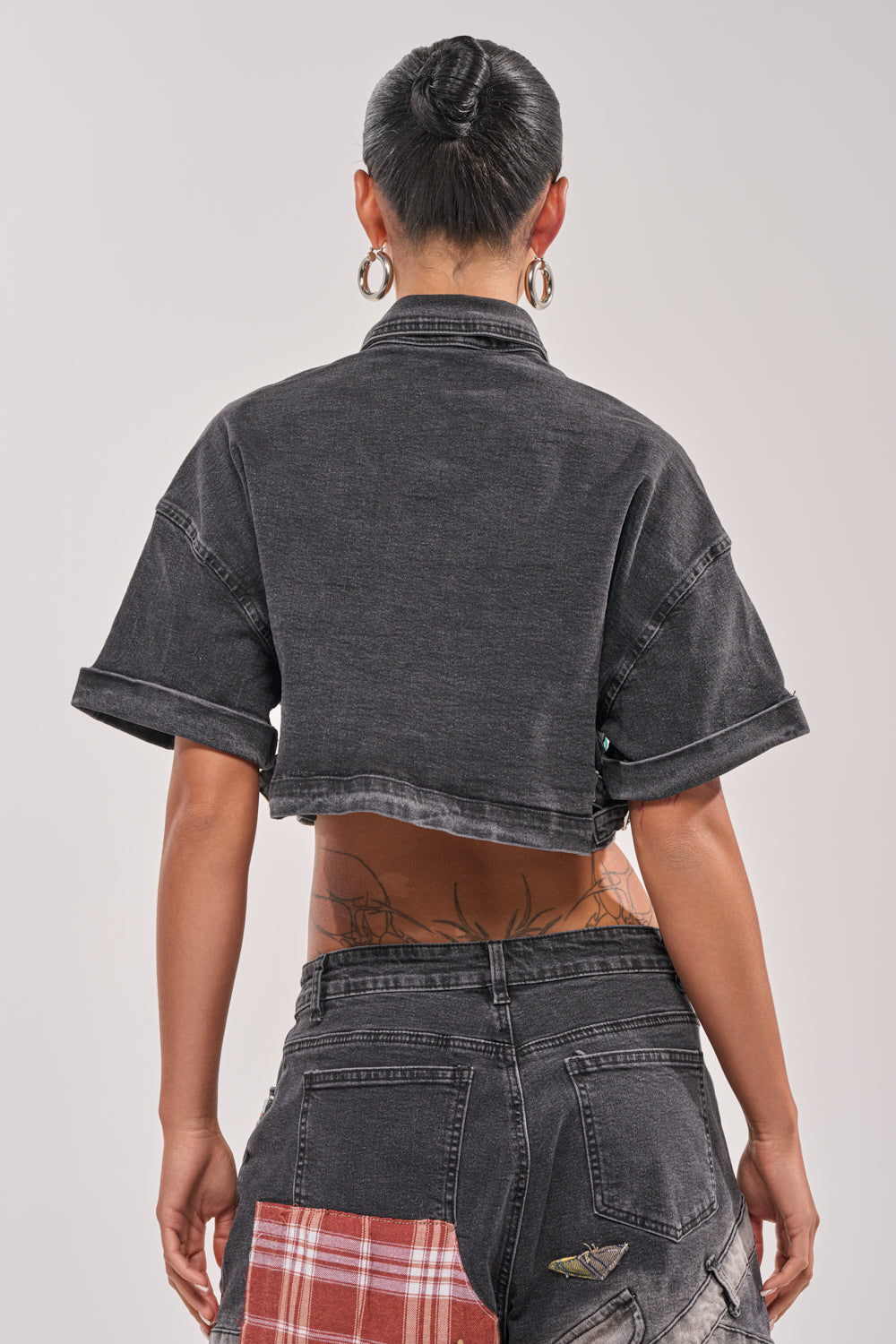 A person stands facing away, wearing the DREA MIXED PLAID CROP SHORT SLEEVE BUTTON DOWN and black denim pants with red plaid patches. With a bun, hoop earrings, and visible back tattoos, they exude effortless street style. 3