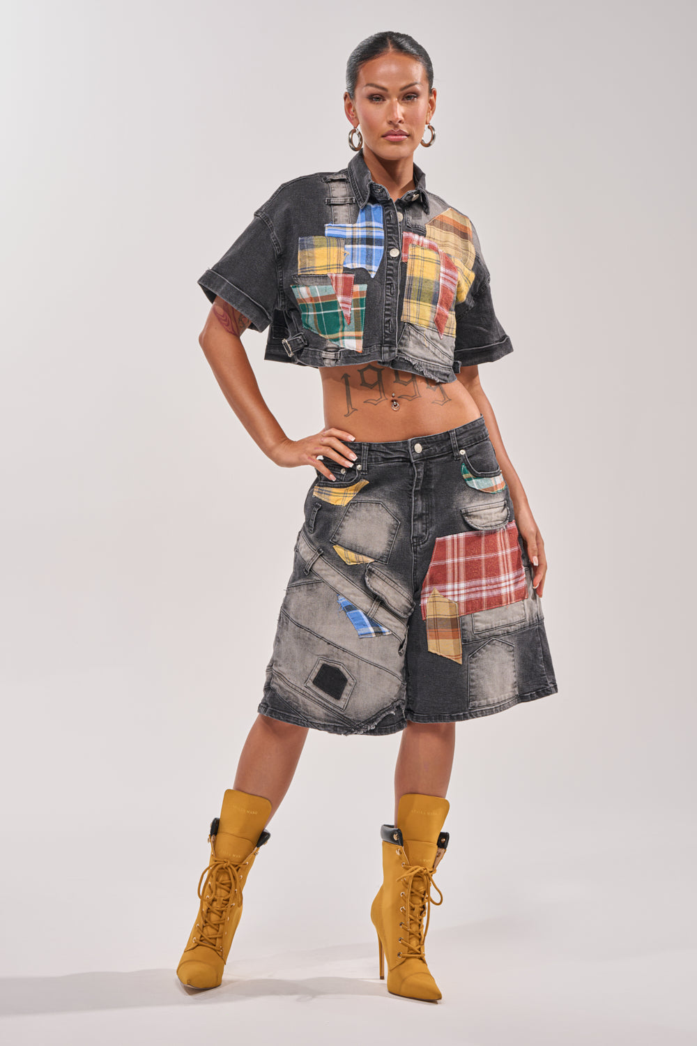 A woman poses confidently in the DREA MIXED PLAID CROP SHORT SLEEVE BUTTON DOWN paired with black denim shorts featuring colorful plaid patches, mustard yellow lace-up boots, and a stomach tattoo. 5