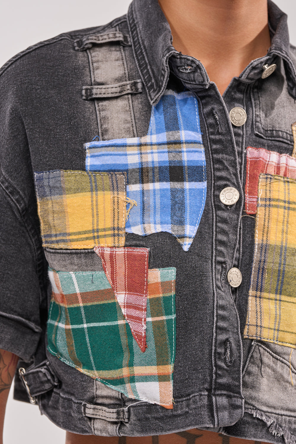 A close-up of the DREA MIXED PLAID CROP SHORT SLEEVE BUTTON DOWN, a black denim shirt with colorful plaid patches, silver buttons, visible stitching, and worn by a medium-skinned model in street-style fashion. 6