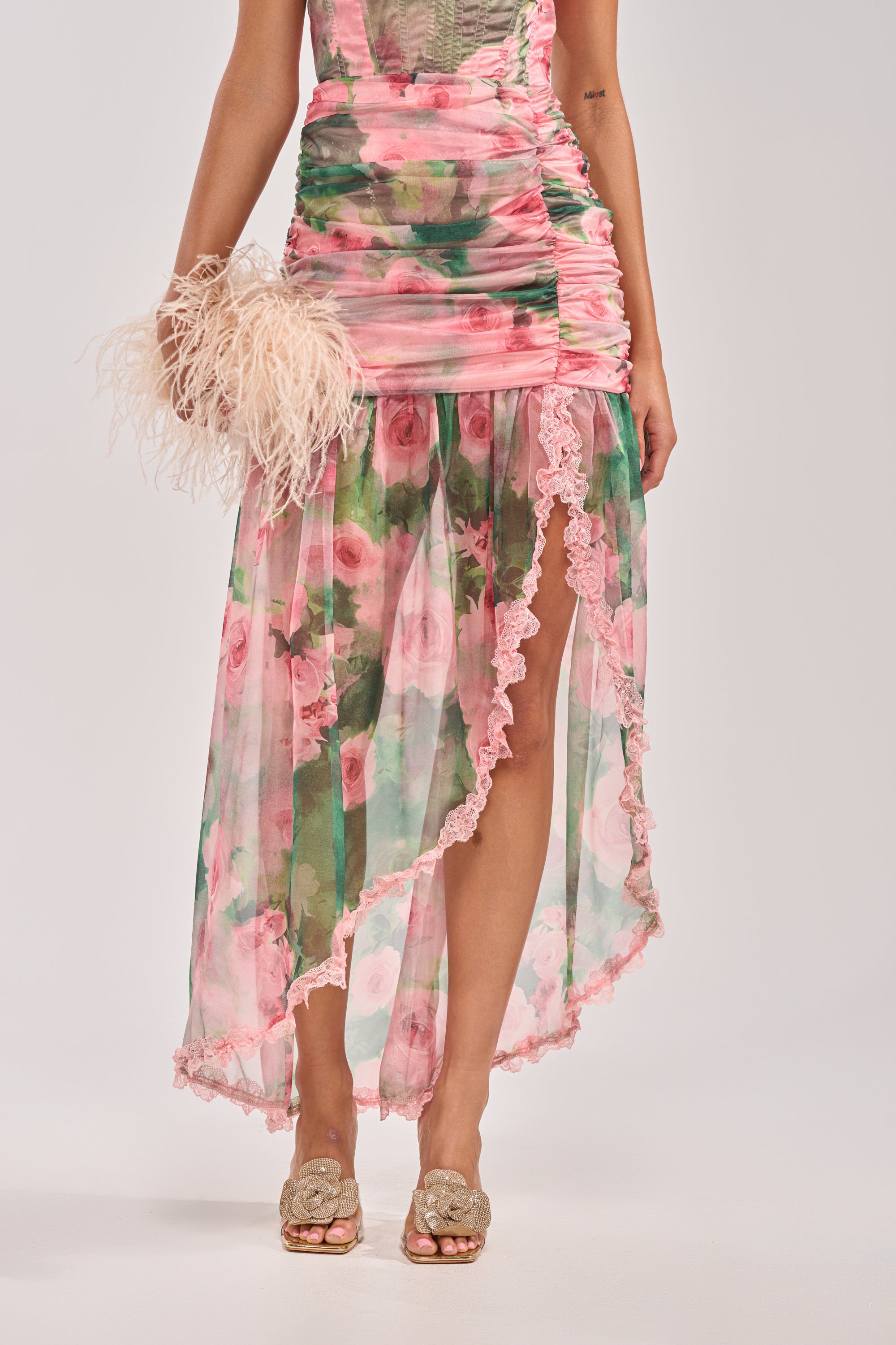 Wearing the DREAMING FLORAL MAXI SKIRT, a sheer pastel high-low skirt with lace trim and high-rise fit, paired with a feathery clutch and gold embellished heels, shown from shoulders down. 0