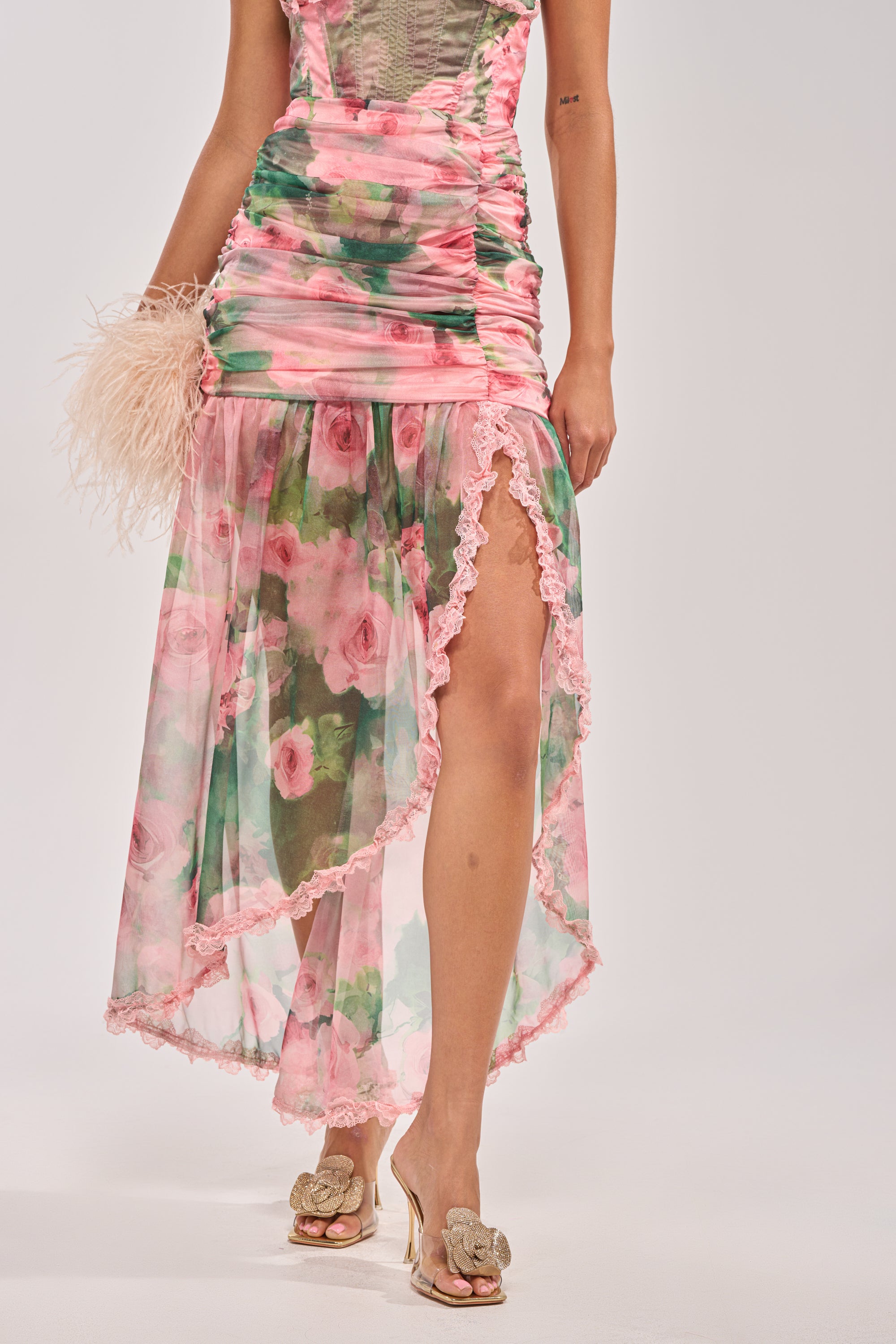 Wearing the DREAMING FLORAL MAXI SKIRT with a pink and green floral print, ruched waist, lace trim, and high slit, she holds a feathered clutch and gold heeled sandals against a plain light background. 1
