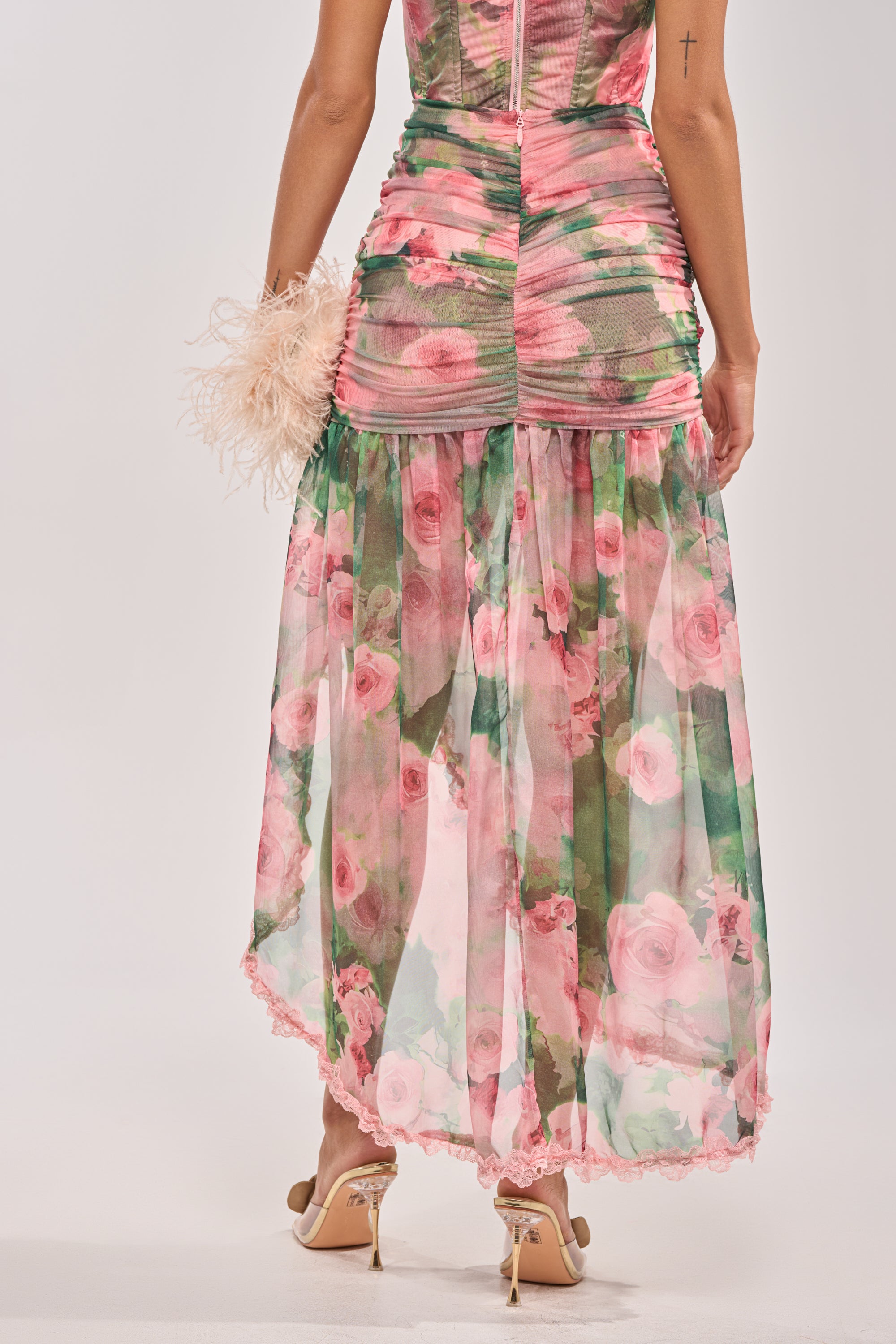 A woman stands facing away, wearing the DREAMING FLORAL MAXI SKIRT—a pink floral pastel chiffon maxi skirt—paired with a ruched top, feathered clutch, and beige high heels. 3