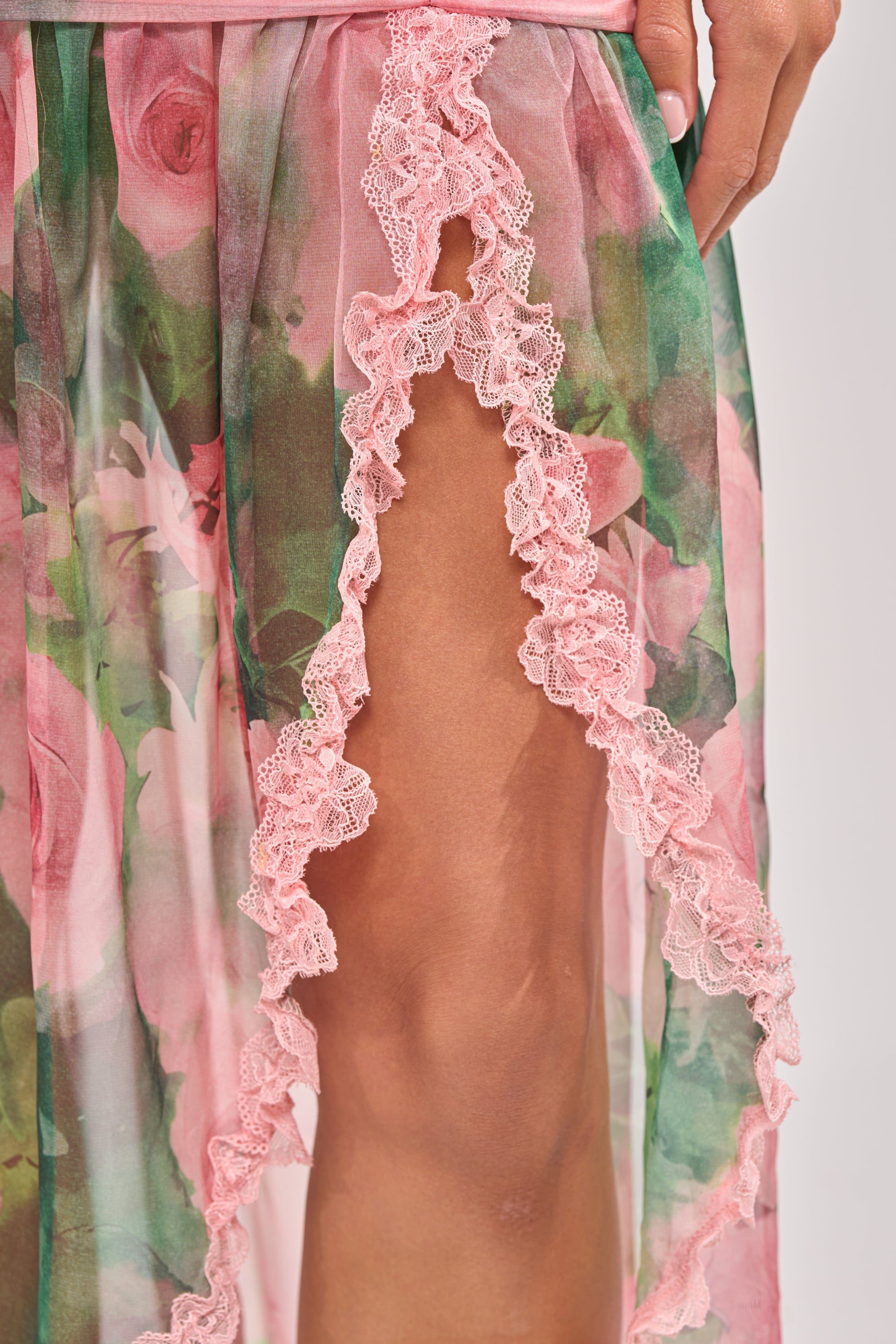 Close-up of a hand on a thigh, wearing the DREAMING FLORAL MAXI SKIRT with chiffon fabric, pastel pink and green rose print, pink lace trim, high slit, and ruched waist that reveals part of the leg. 7