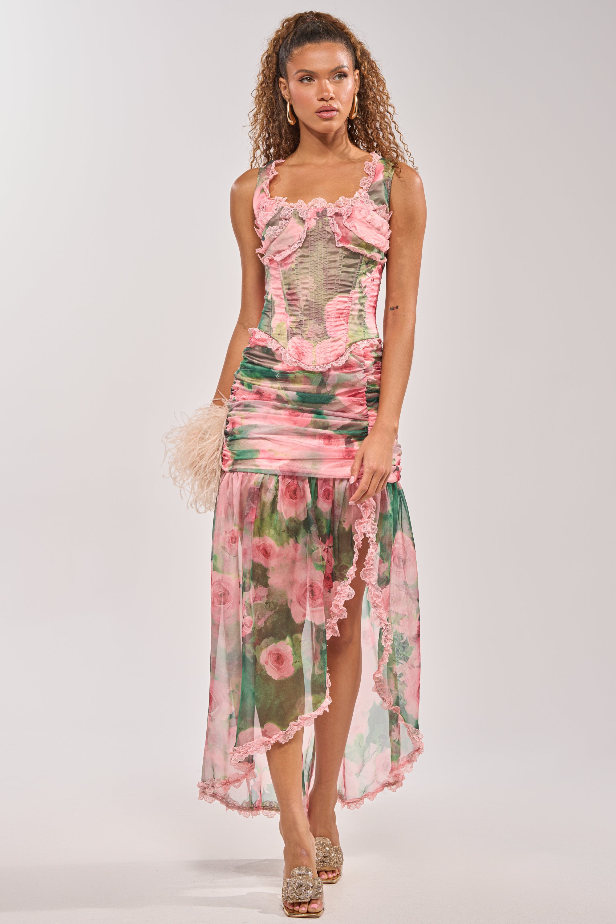 A woman with curly hair wears the DREAMING FLORAL CORSET—a pink and green floral dress with ruffles, a fitted bodice, and a sheer asymmetrical skirt—while holding a feathered clutch and sparkly heels against a white background. 6