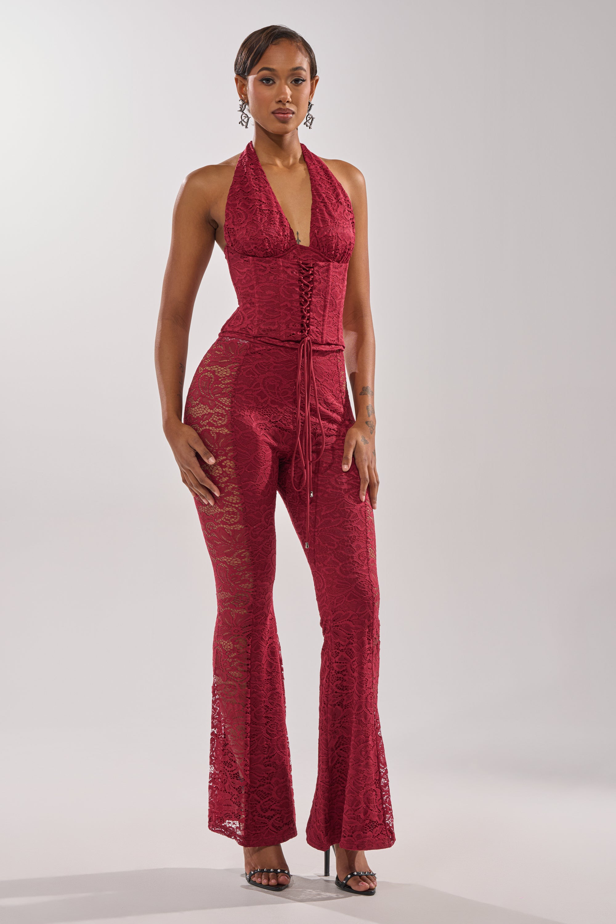 A woman with short hair and drop earrings poses against a plain background wearing the LOVE BOMB LACE PANT—a sleeveless, deep red lace halter jumpsuit with flared legs—paired with open-toe heels.