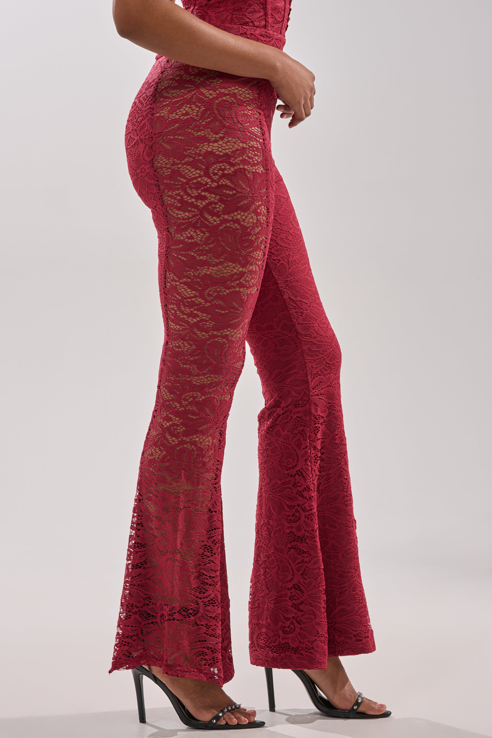A woman stands sideways against a plain light background wearing the LOVE BOMB LACE PANT in red floral lace paired with black high-heeled sandals.