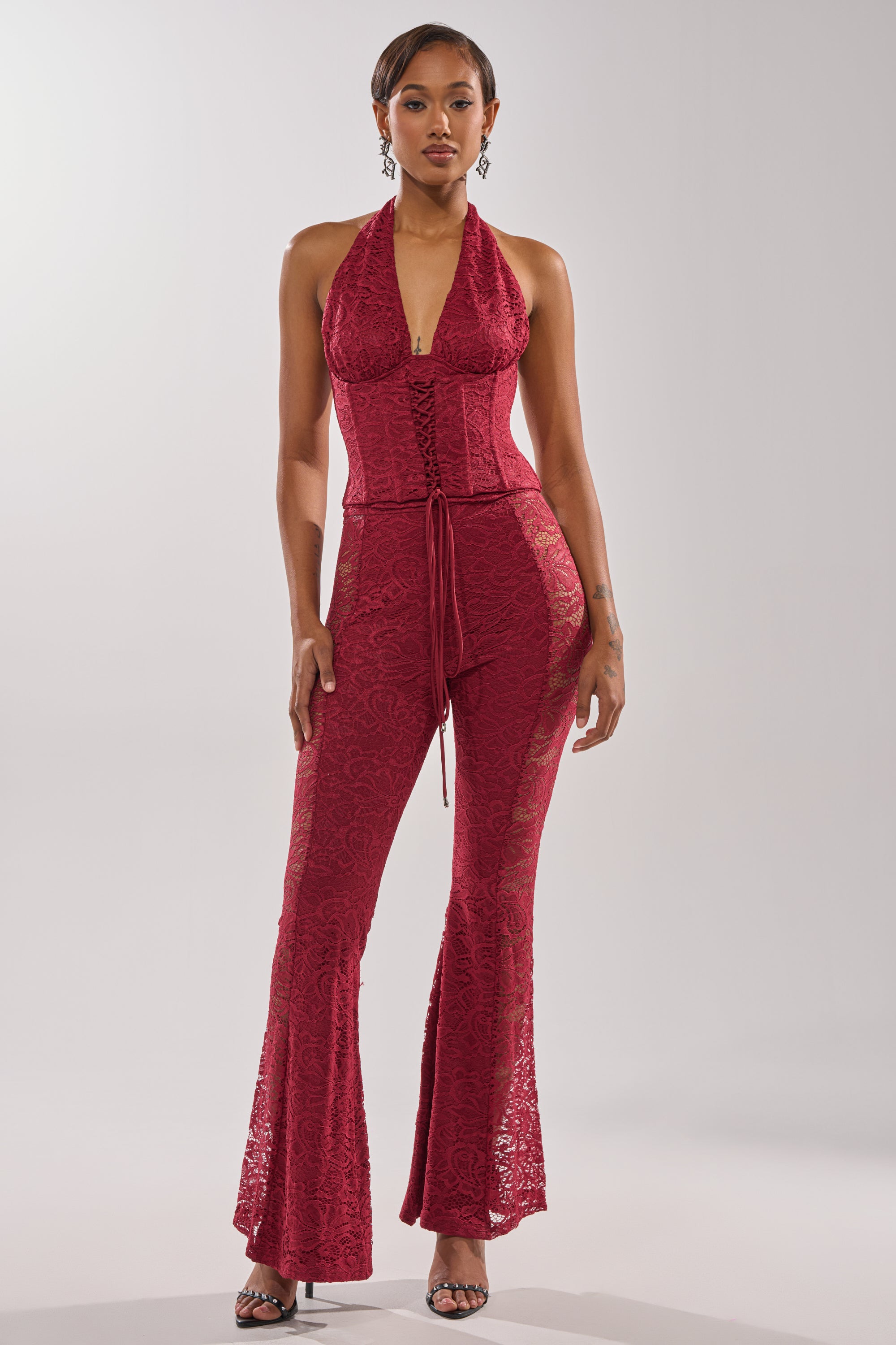 A woman models the LOVE BOMB LACE PANT, a deep red halter jumpsuit with a corset bodice, high-rise flared floral lace pants, and waist tie. She styles it with open-toed heels and slicked-back short hair.