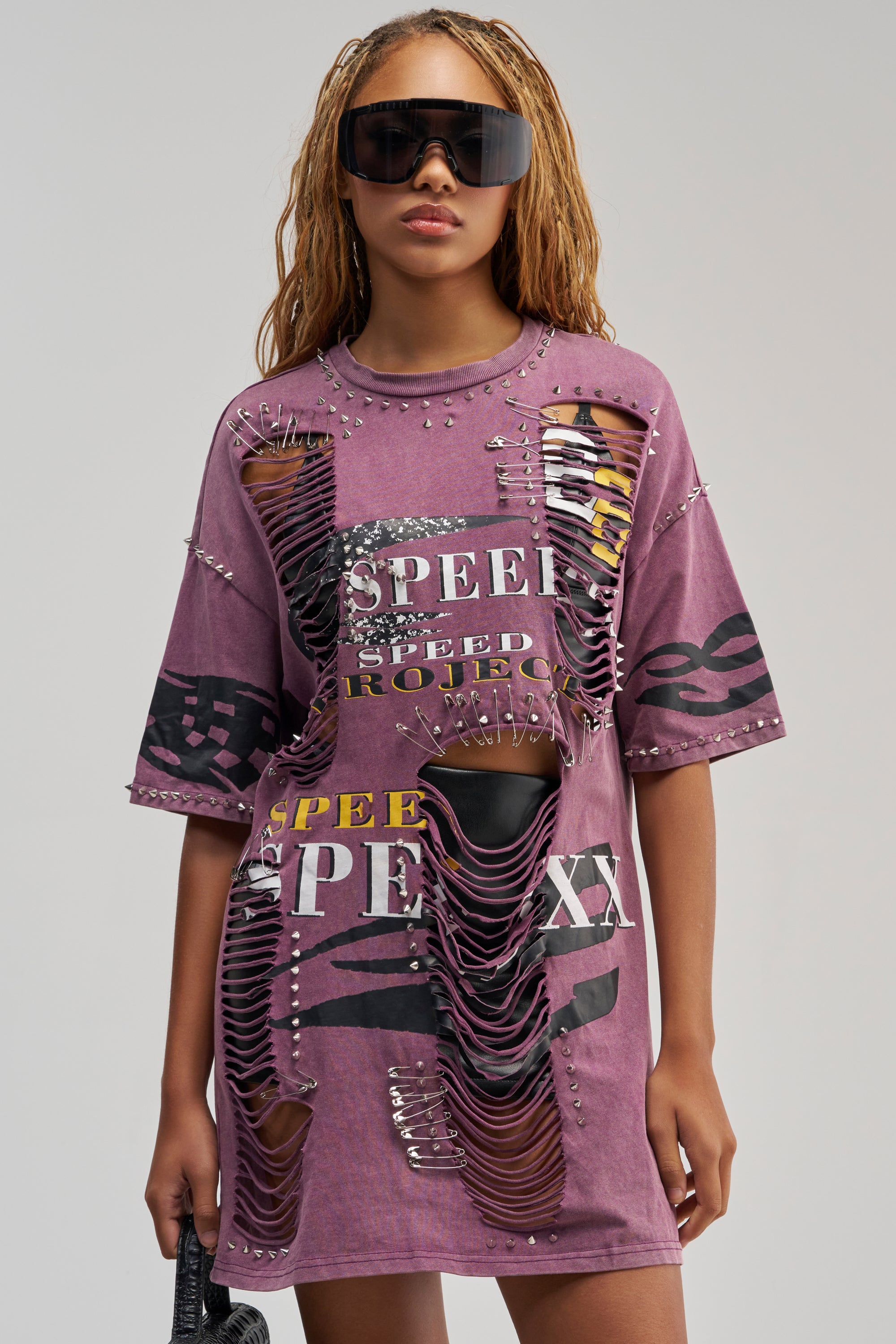 A woman with long braids stands against a plain background, wearing the NEVER TOO MUCH DISTRESSED OVERSIZED TEE IN PLUM—an edgy streetwear dress featuring “SPEED” text, zebra and leopard prints, studs, safety pin accents, and shredded details. She completes her look with oversized sunglasses for a bold statement.
