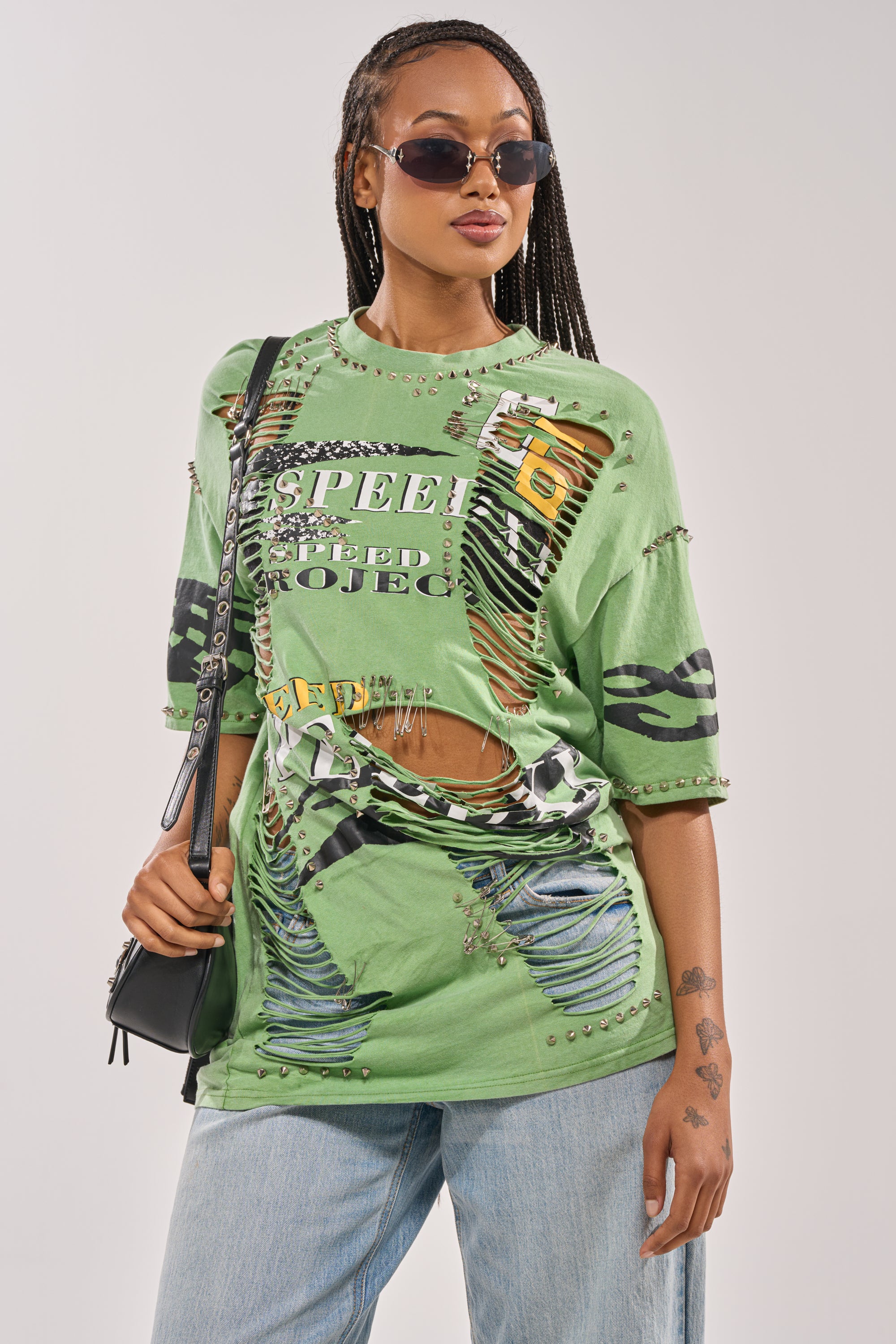 A stylish woman with braided hair wears sunglasses, light blue jeans, a black shoulder bag, and the NEVER TOO MUCH DISTRESSED OVERSIZED TEE, posing confidently against a plain background. 2