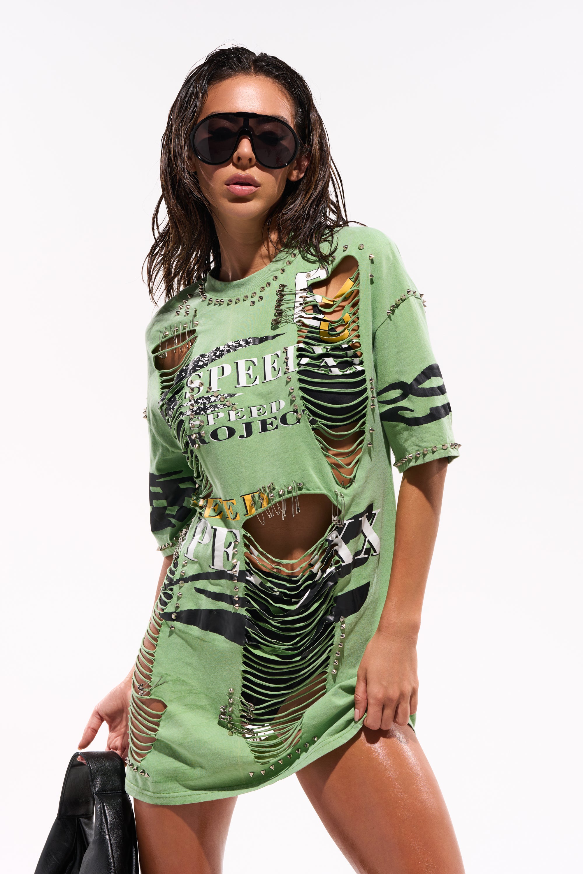 A woman stands confidently in large black sunglasses and the NEVER TOO MUCH DISTRESSED OVERSIZED TEE with safety pin accents and graphic text, holding a black jacket for a bold streetwear look. 0