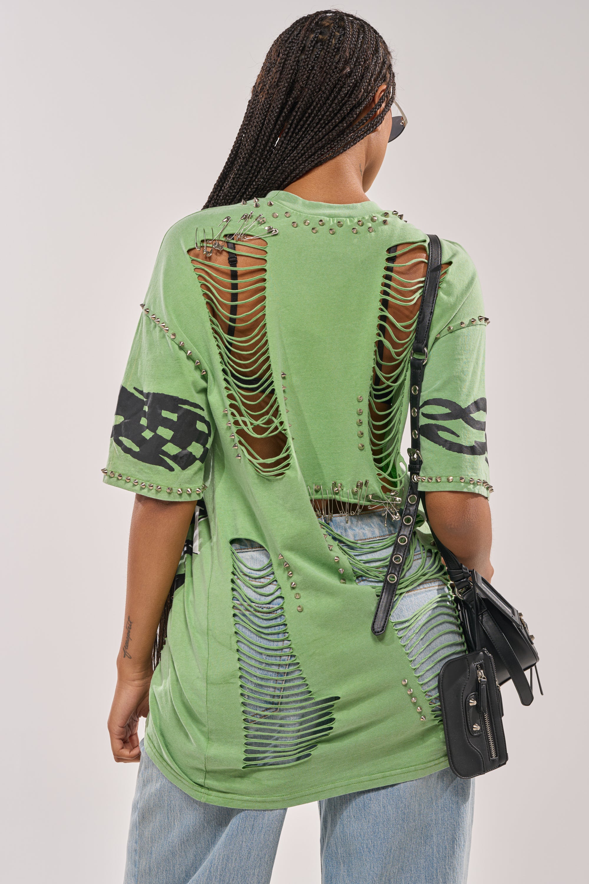 A person with braided hair rocks streetwear in the NEVER TOO MUCH DISTRESSED OVERSIZED TEE—green, oversized, with shredded and ripped details, metal studs, and black sleeve designs—paired with a black crossbody bag and light blue jeans. 6