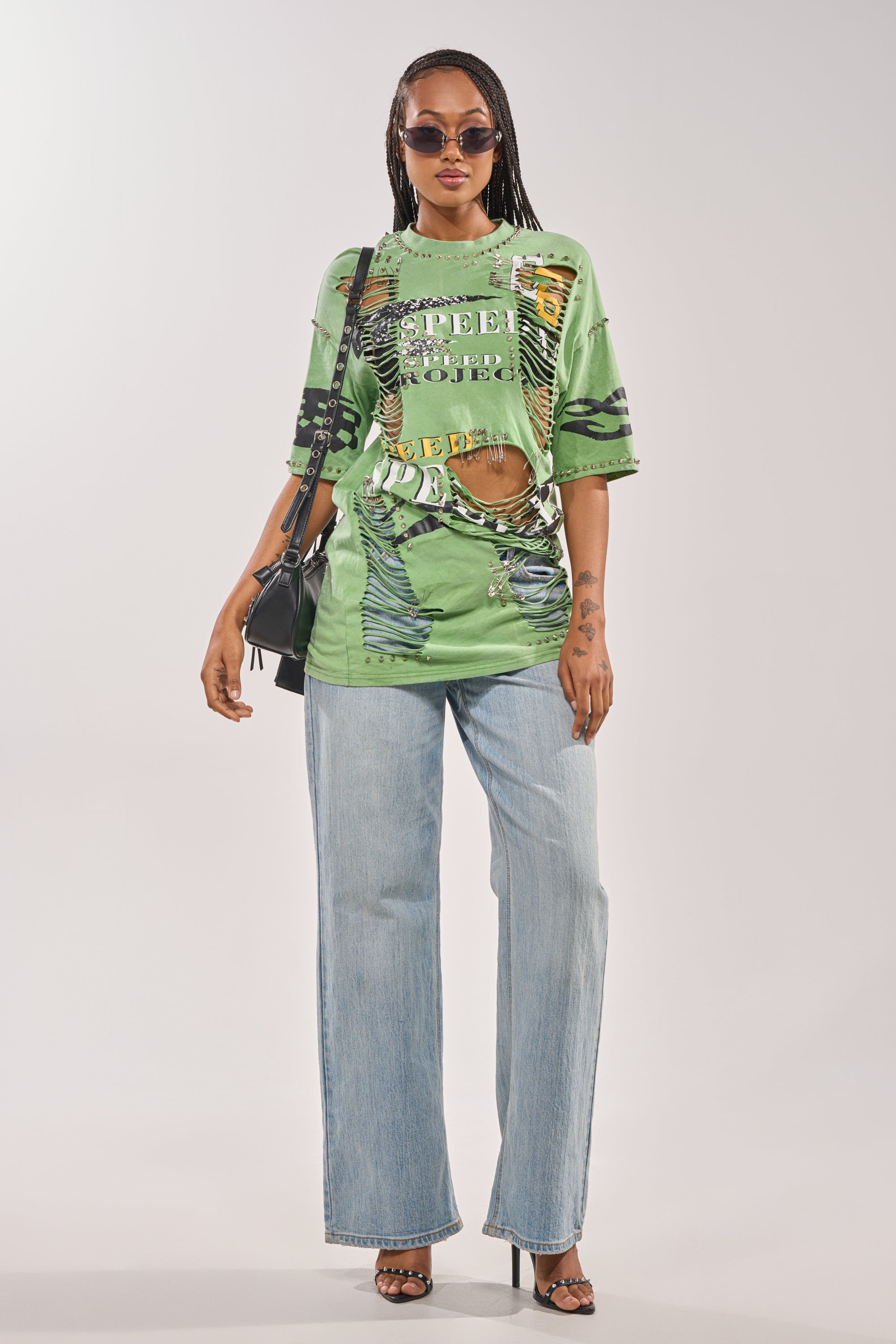 A woman wears the NEVER TOO MUCH DISTRESSED OVERSIZED TEE, styled with sunglasses, light blue wide-leg jeans, black heeled sandals, and a black shoulder bag, standing against a plain background. 7