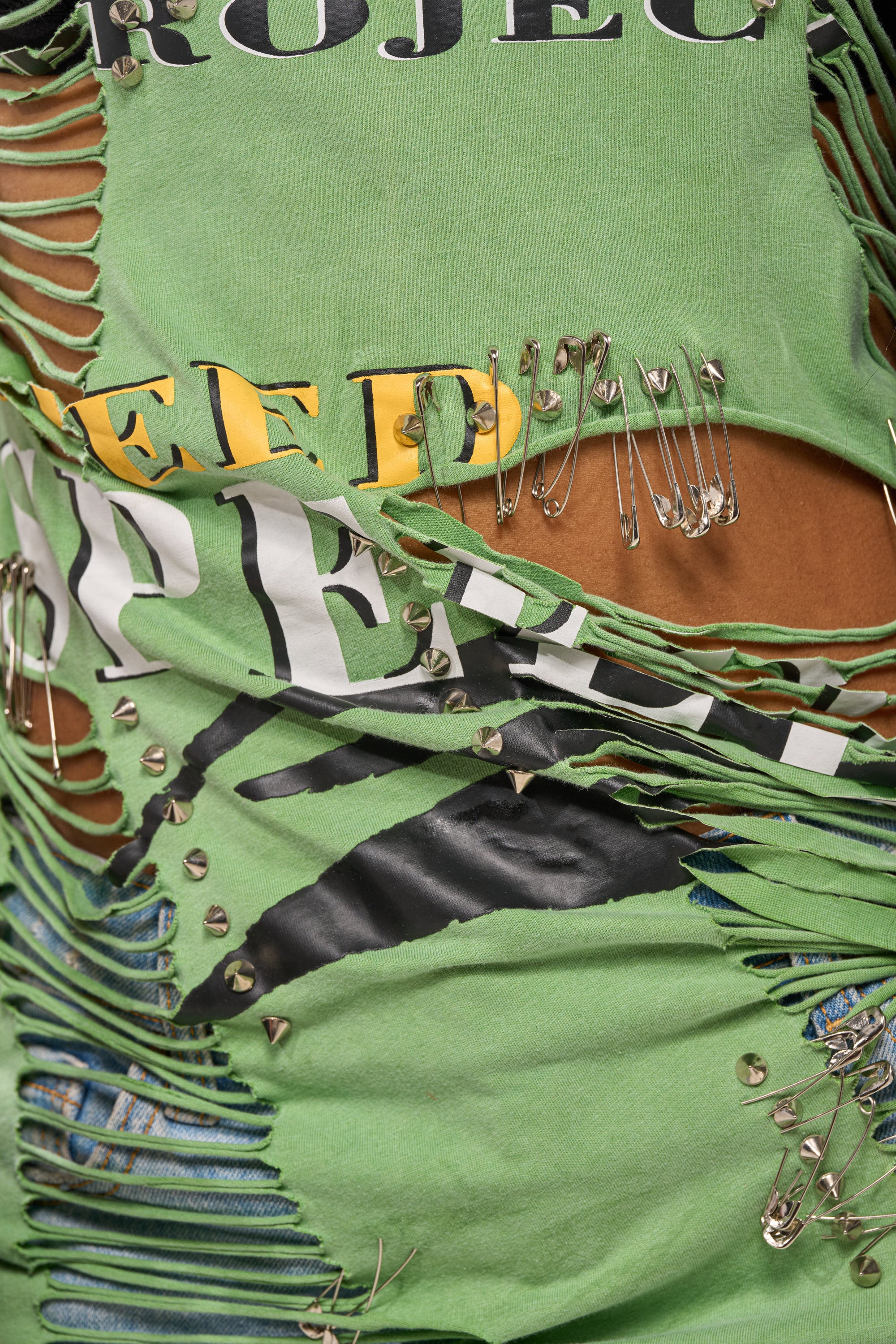 The NEVER TOO MUCH DISTRESSED OVERSIZED TEE features shredded green fabric, brown underlayers, safety pin accents, metal studs, and multicolored printed text—a bold choice for any streetwear style. 8