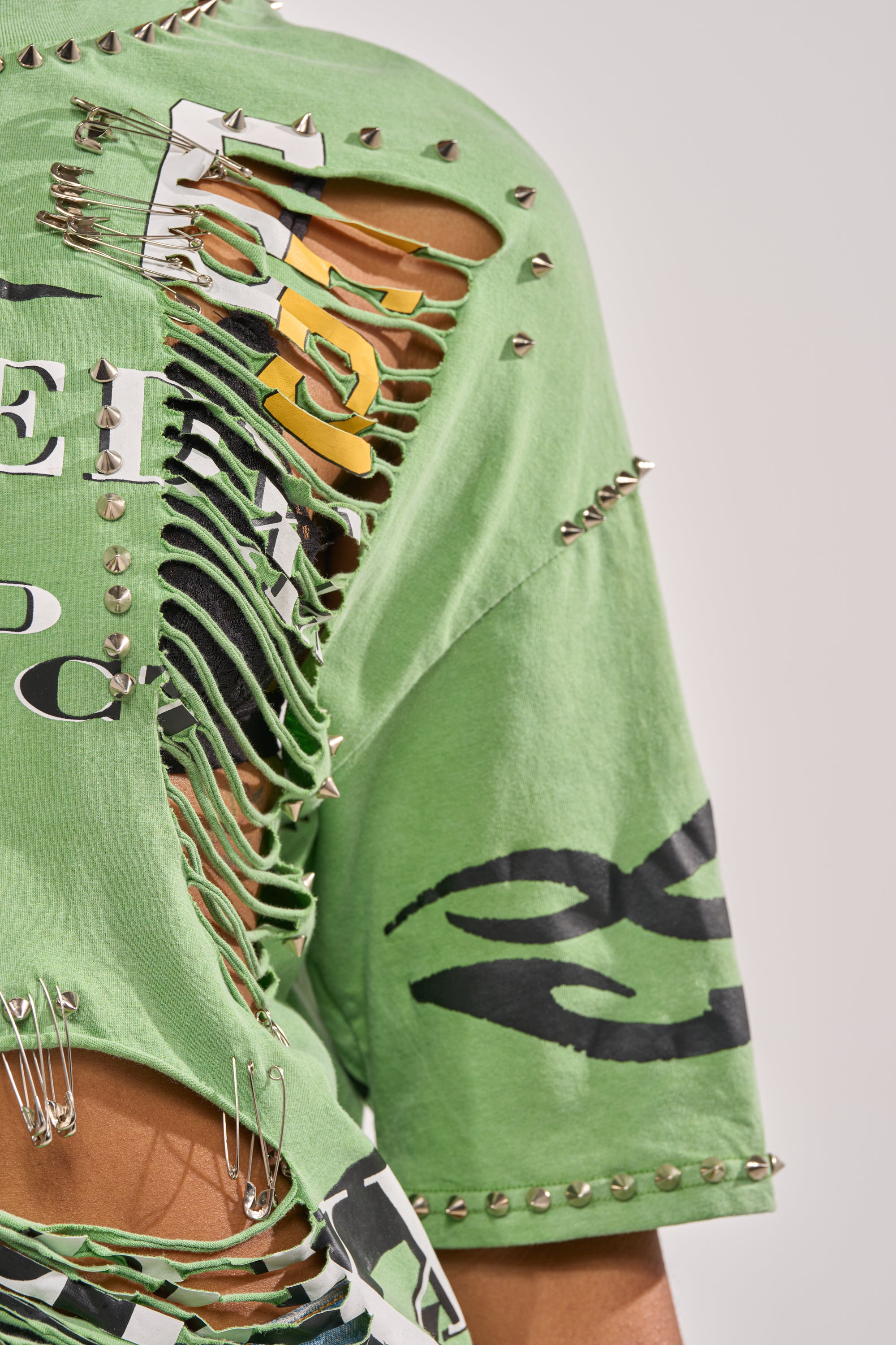 Close-up of the NEVER TOO MUCH DISTRESSED OVERSIZED TEE in green, featuring safety pin details, silver studs, black-and-white prints, and ripped sections revealing layered fabric for a bold streetwear vibe. 9