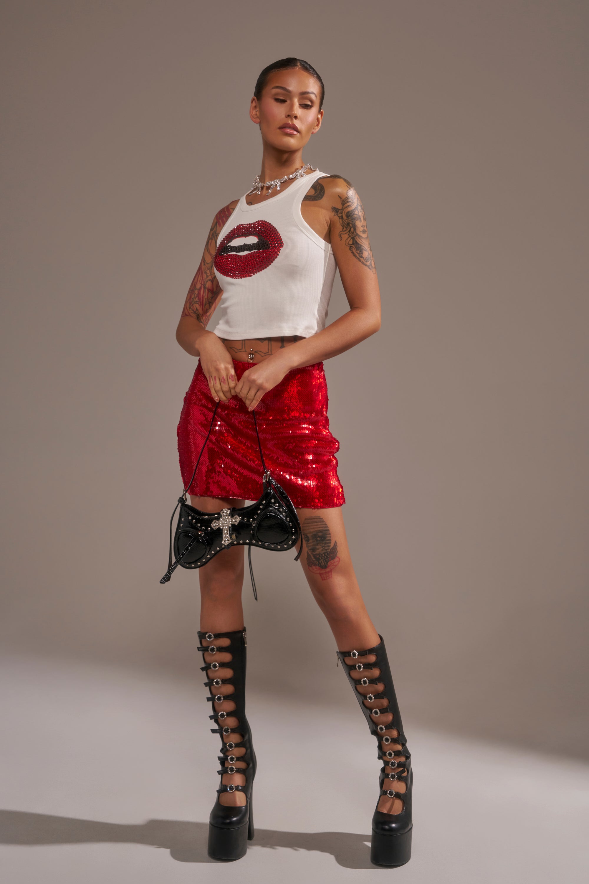 A woman exudes confidence in the KISS ME BABY SEQUIN MINI SKIRT, styled with a white tank featuring a red lip graphic, black lace-up platform boots, a studded handbag, visible tattoos, and a sleek hairstyle.