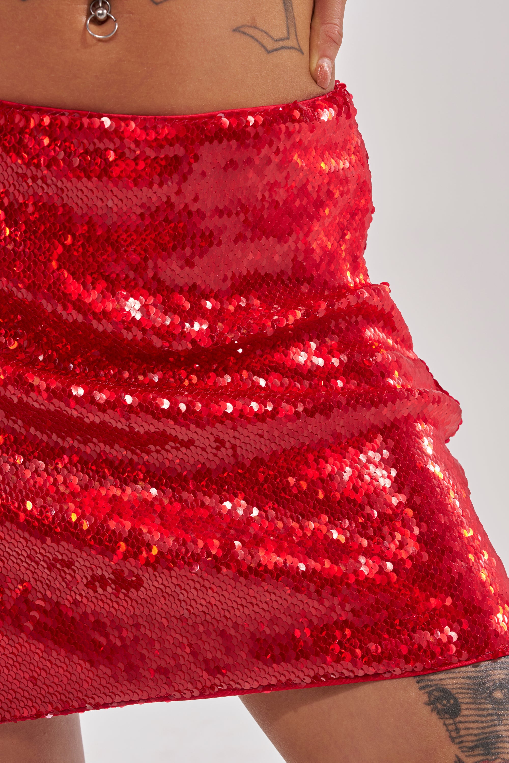 A person wearing the KISS ME BABY SEQUIN MINI SKIRT in shiny red stands with one hand on their waist, showing tattoos on their hip and thigh against a plain, light-colored background.