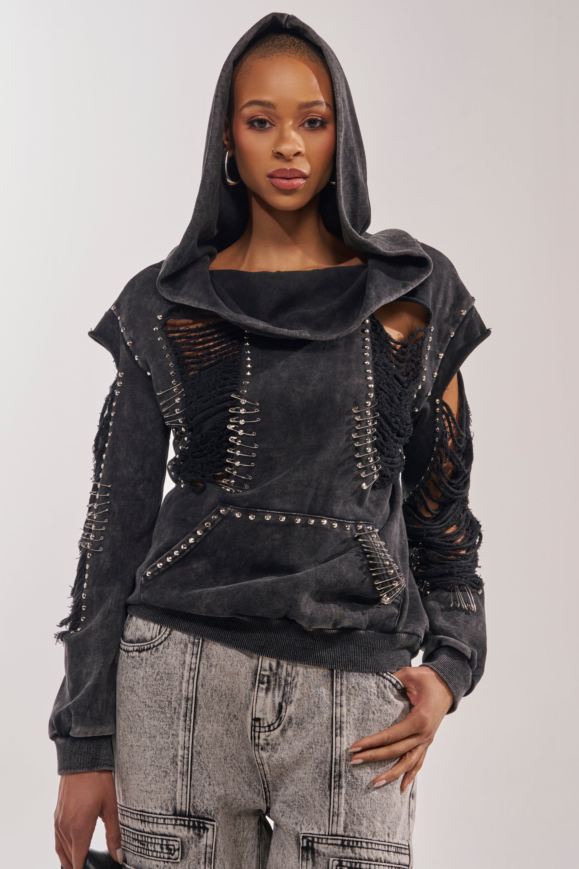 A woman models the ALL TORN UP DISTRESSED OFF THE SHOULDER SWEATSHIRT, a black top with metal studs and ripped details, styled with high-waisted, light-wash jeans featuring seam accents for an edgy streetwear look. 0