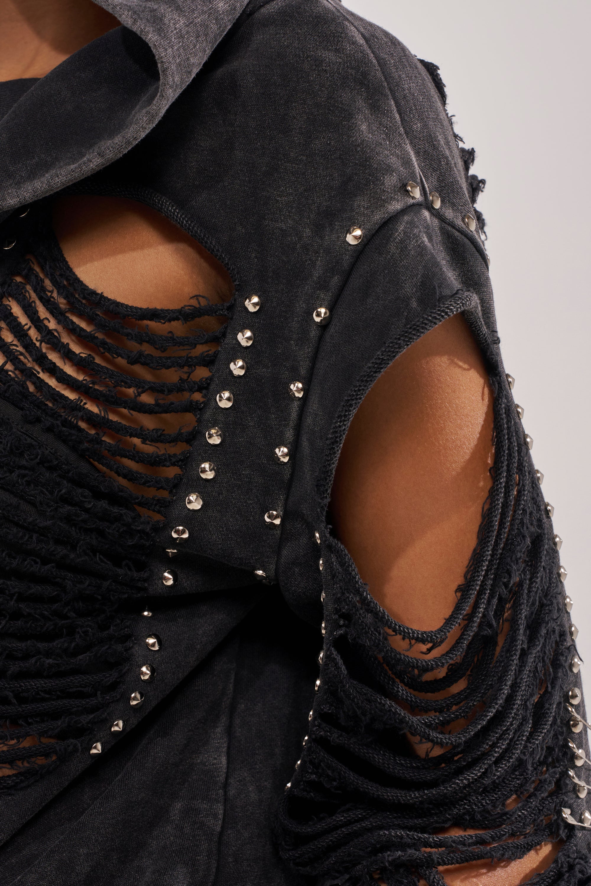 Close-up of the ALL TORN UP DISTRESSED OFF THE SHOULDER SWEATSHIRT, featuring black shredded fabric and silver studs on the edges. The punk-inspired design is shown worn off one shoulder by a person with medium-brown skin. 9