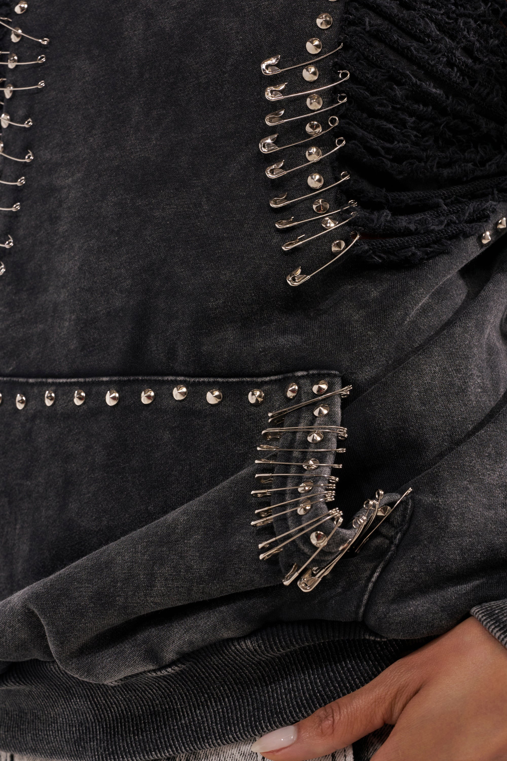 Close-up of the ALL TORN UP DISTRESSED OFF THE SHOULDER SWEATSHIRT, detailed with black fabric, bold silver safety pin embellishments, and edgy studded accents—a perfect statement piece for punk-inspired streetwear lovers. A manicured hand adds extra flair. 10