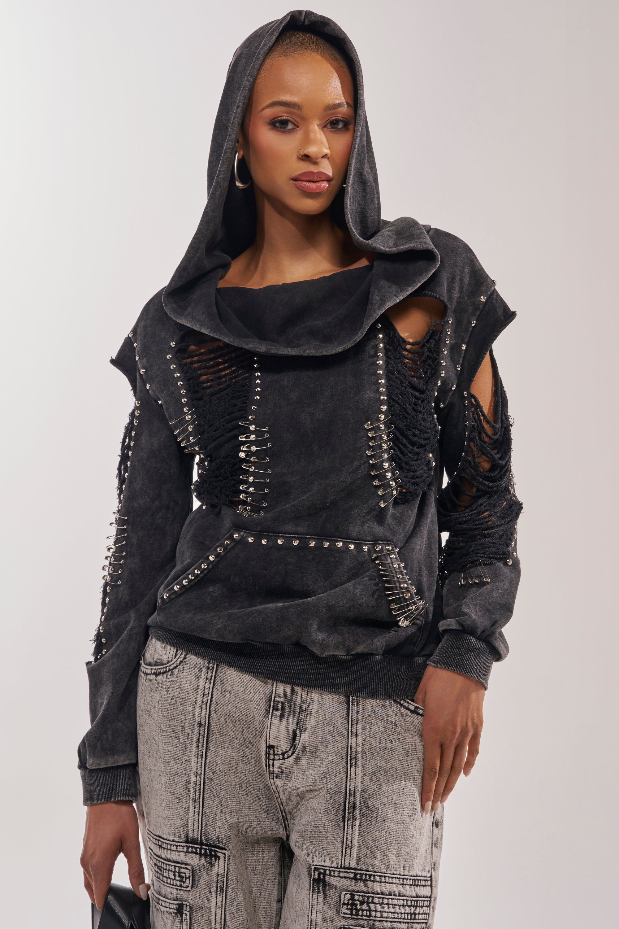 A woman faces forward in a punk streetwear style, pulling up the hood of an ALL TORN UP Distressed Off The Shoulder Sweatshirt with tears and studs, styled with light gray patterned pants and a black bag. 1