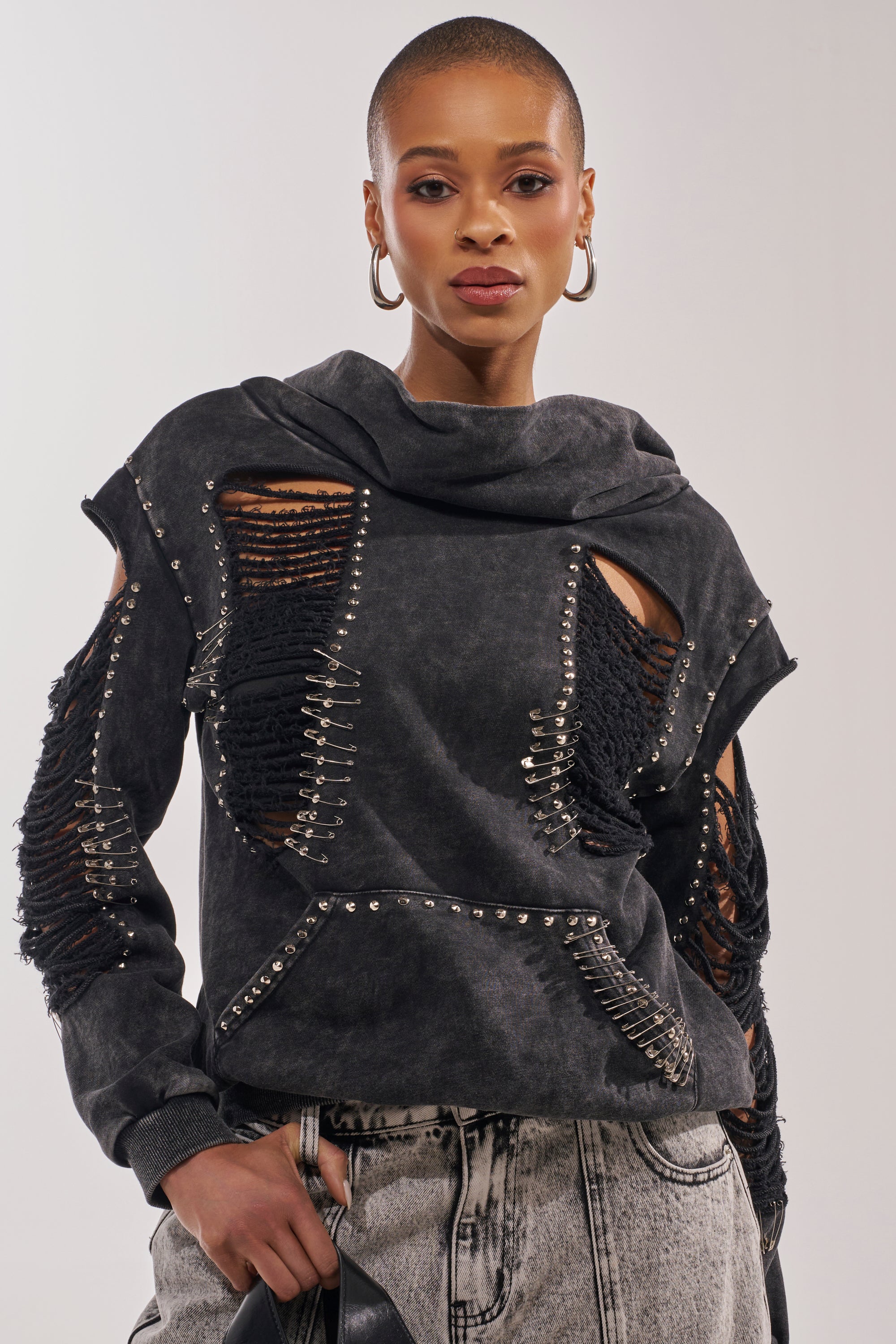 A person with a shaved head wears large hoop earrings, light gray jeans, and the ALL TORN UP DISTRESSED OFF THE SHOULDER SWEATSHIRT with shredded details, confidently posing in punk-inspired streetwear against a neutral background. 2