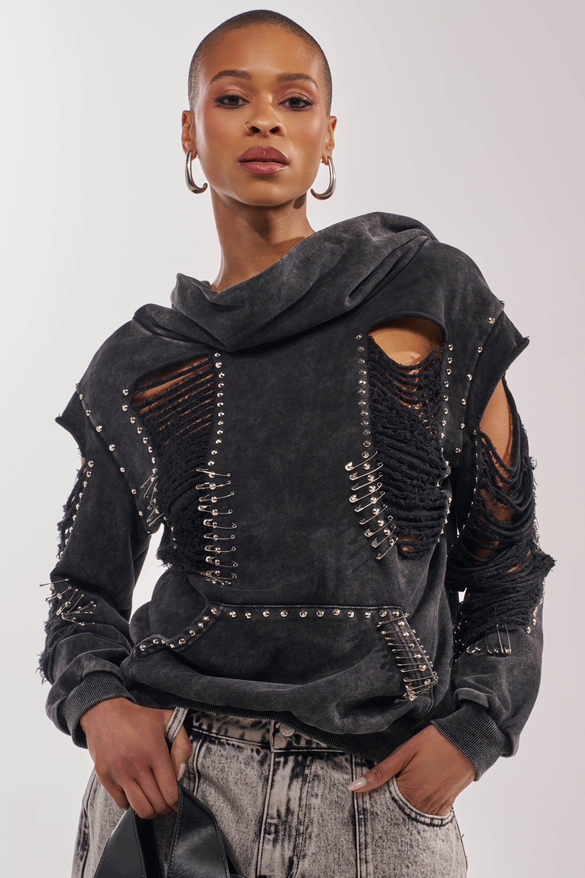 A woman rocks punk streetwear in the ALL TORN UP DISTRESSED OFF THE SHOULDER SWEATSHIRT with rips and metal studs, paired with acid-wash jeans. She wears hoop earrings, has closely-cropped hair, and holds a black item in one hand. 3