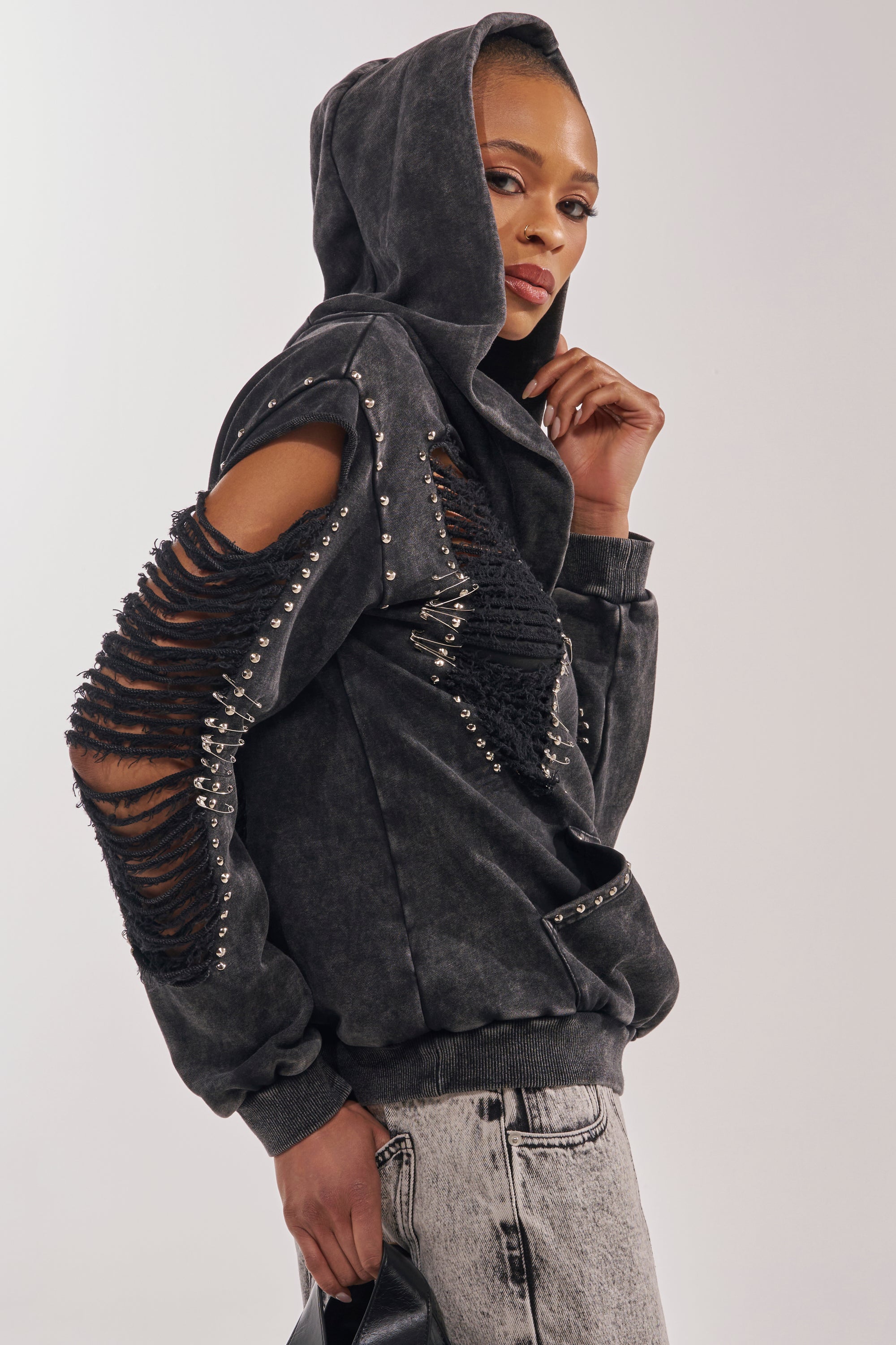 A stylish individual rocks the ALL TORN UP DISTRESSED OFF THE SHOULDER SWEATSHIRT with ripped sleeves and hood up, light-wash jeans, and a black bag, confidently posing against a plain background in punk-inspired streetwear. 4