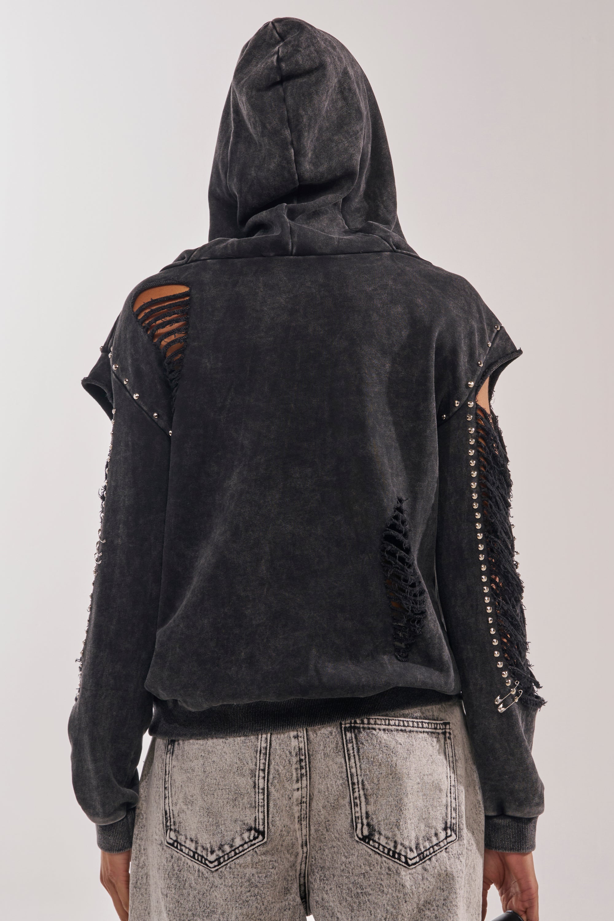 Someone wears the ALL TORN UP DISTRESSED OFF THE SHOULDER SWEATSHIRT with metal-studded sleeves and light gray acid-wash jeans, showcasing a punk-inspired streetwear look, seen from behind against a plain background. 5