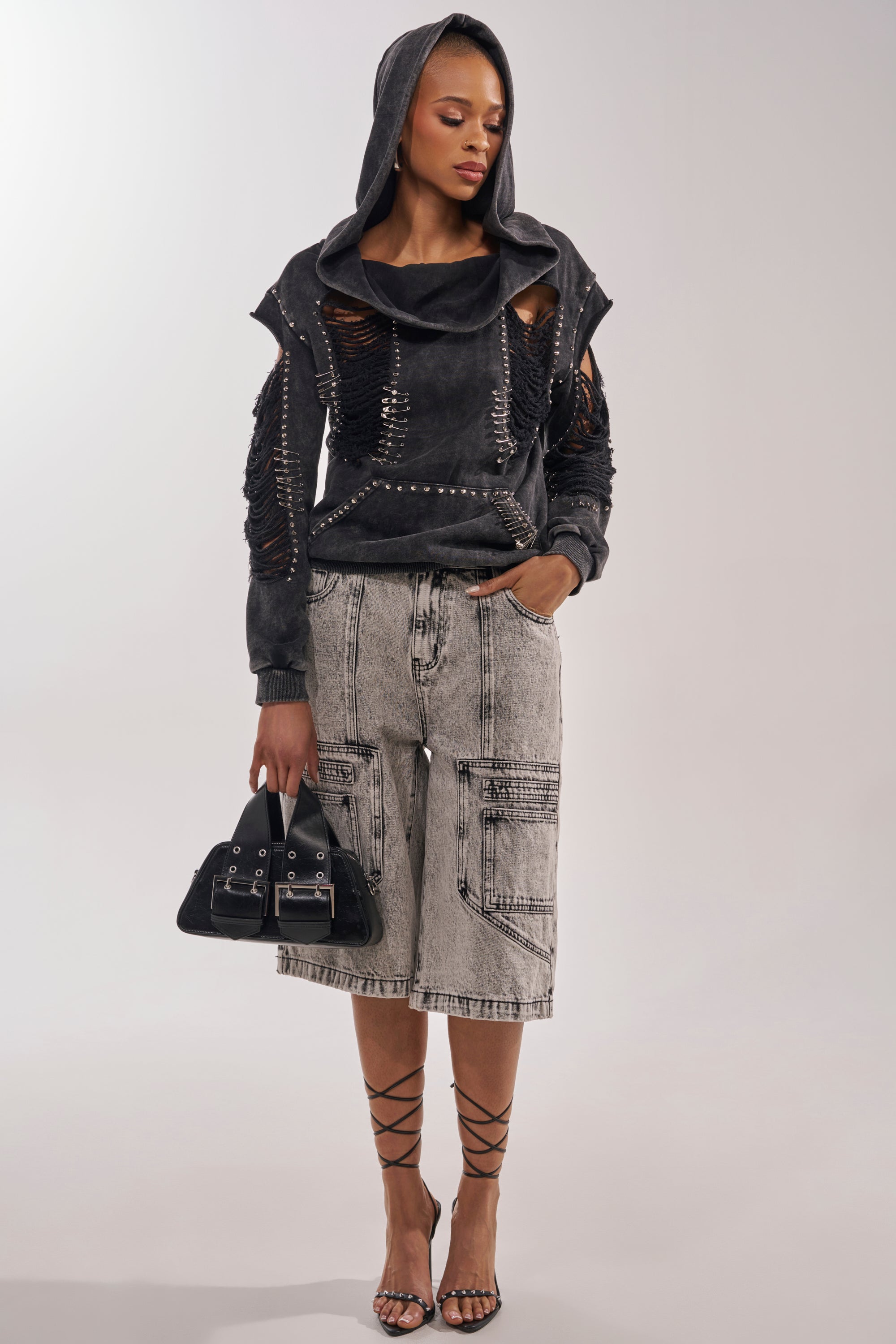 A woman stands against a neutral background wearing the ALL TORN UP DISTRESSED OFF THE SHOULDER SWEATSHIRT with ripped sleeves, light gray knee-length cargo shorts, strappy black heels, and a black handbag. 6