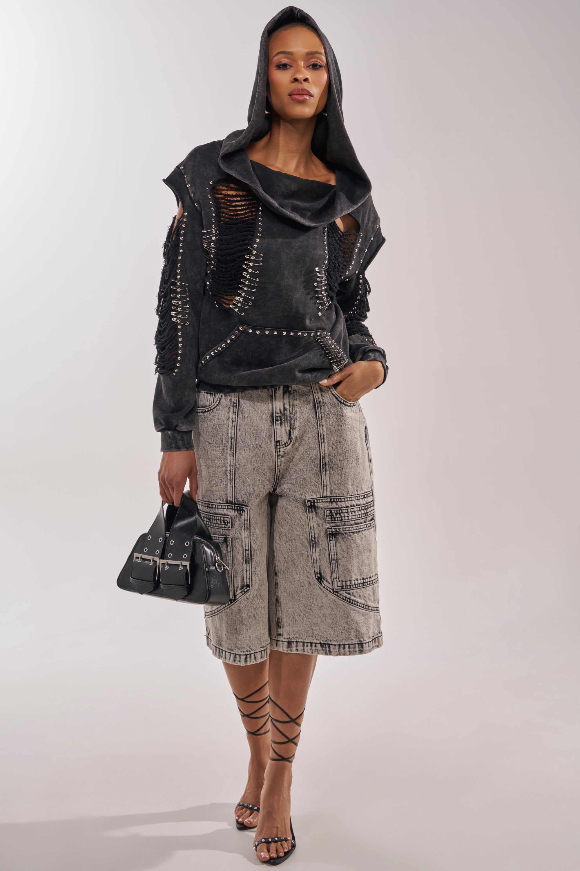 A woman wears the ALL TORN UP DISTRESSED OFF THE SHOULDER SWEATSHIRT, light gray knee-length denim shorts, strappy black heeled sandals, and carries a small black handbag for a modern grunge look. 7