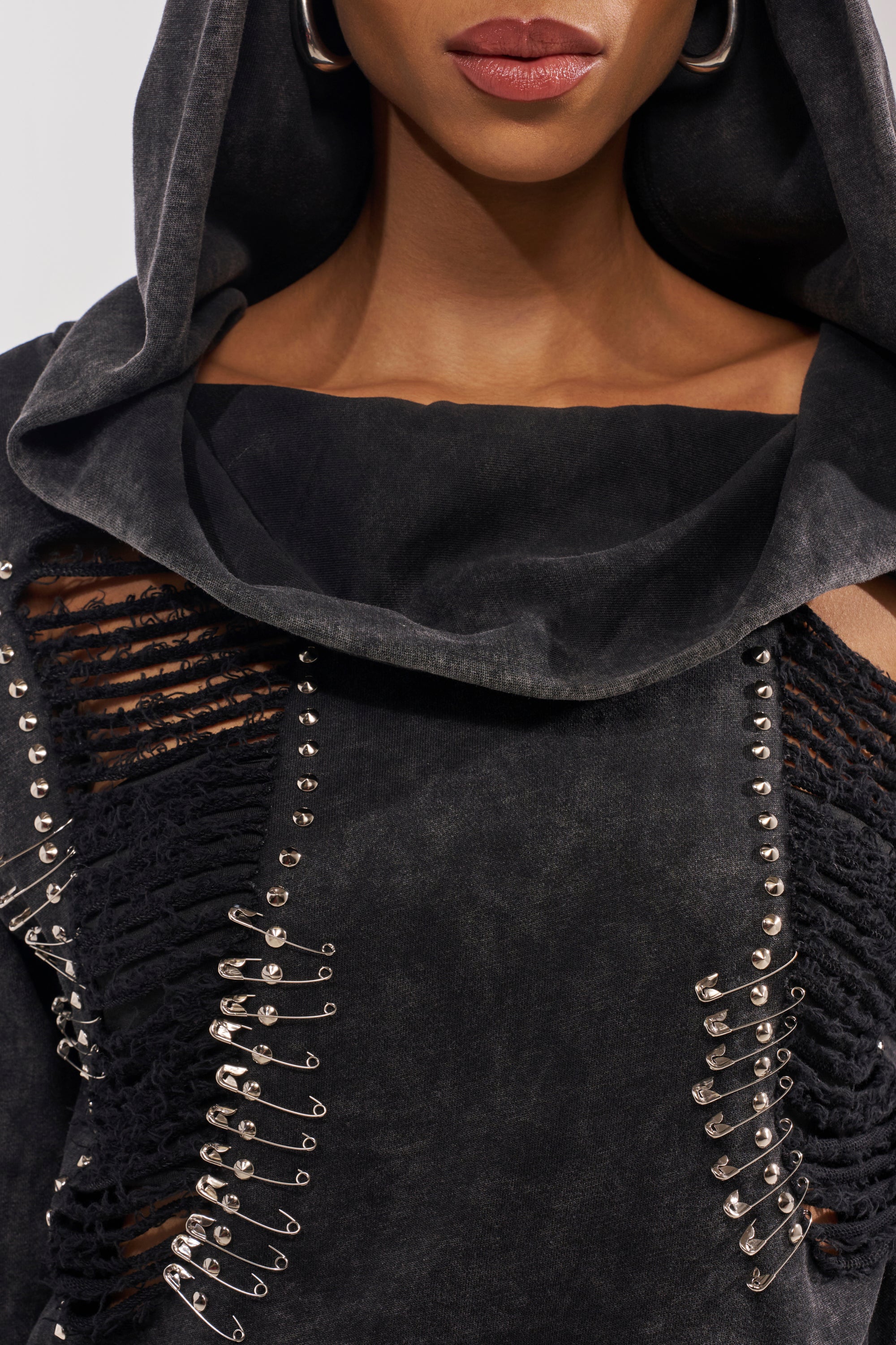 A close-up of a person wearing the ALL TORN UP DISTRESSED OFF THE SHOULDER SWEATSHIRT, featuring black fabric with studs and rows of safety pins—a bold choice for grunge or punk streetwear. Only their lower face and neutral lips are visible. 8