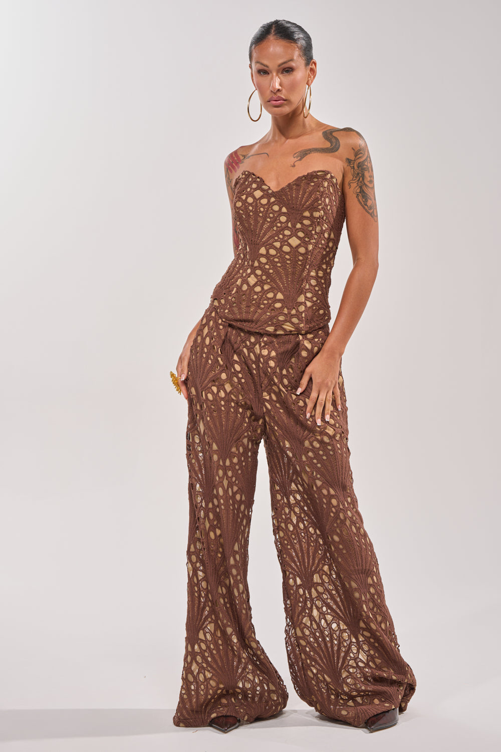 A woman stands confidently against a plain, light background wearing the CLAIRE EYELET PALAZZO PANT, a brown wide-leg pant with an eyelet design. She pairs it with large hoop earrings, has visible tattoos, and her hair is pulled back. 0
