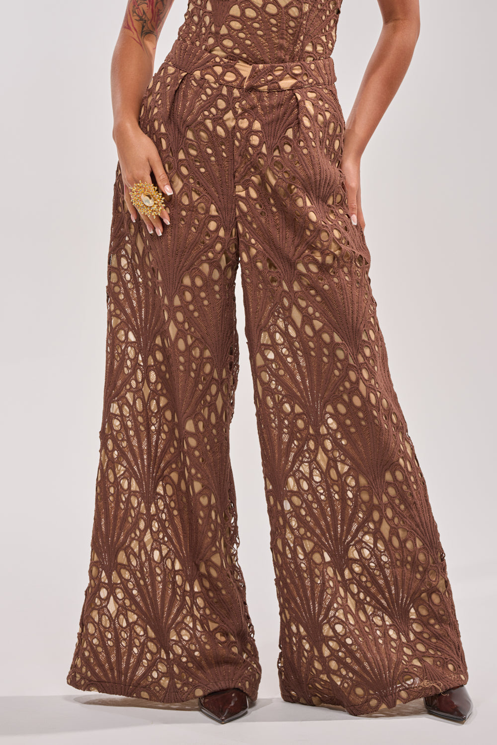 Wearing the CLAIRE EYELET PALAZZO PANT, featuring high-waisted, wide-leg style with a geometric eyelet design over nude lining, this person poses with one hand on their hip and sports a large ring. 1