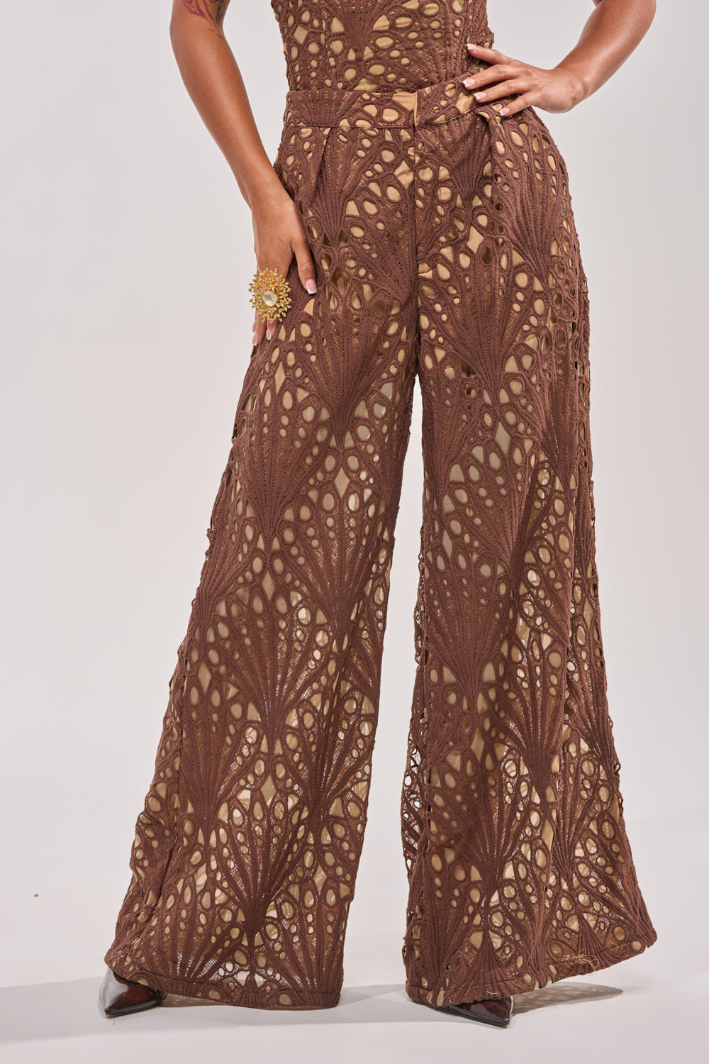 Wearing the CLAIRE EYELET PALAZZO PANT—wide-leg, brown with a geometric eyelet pattern and beige lining—a person poses with one hand on their hip, a bold gold floral ring, and pointed-toe brown shoes. 2