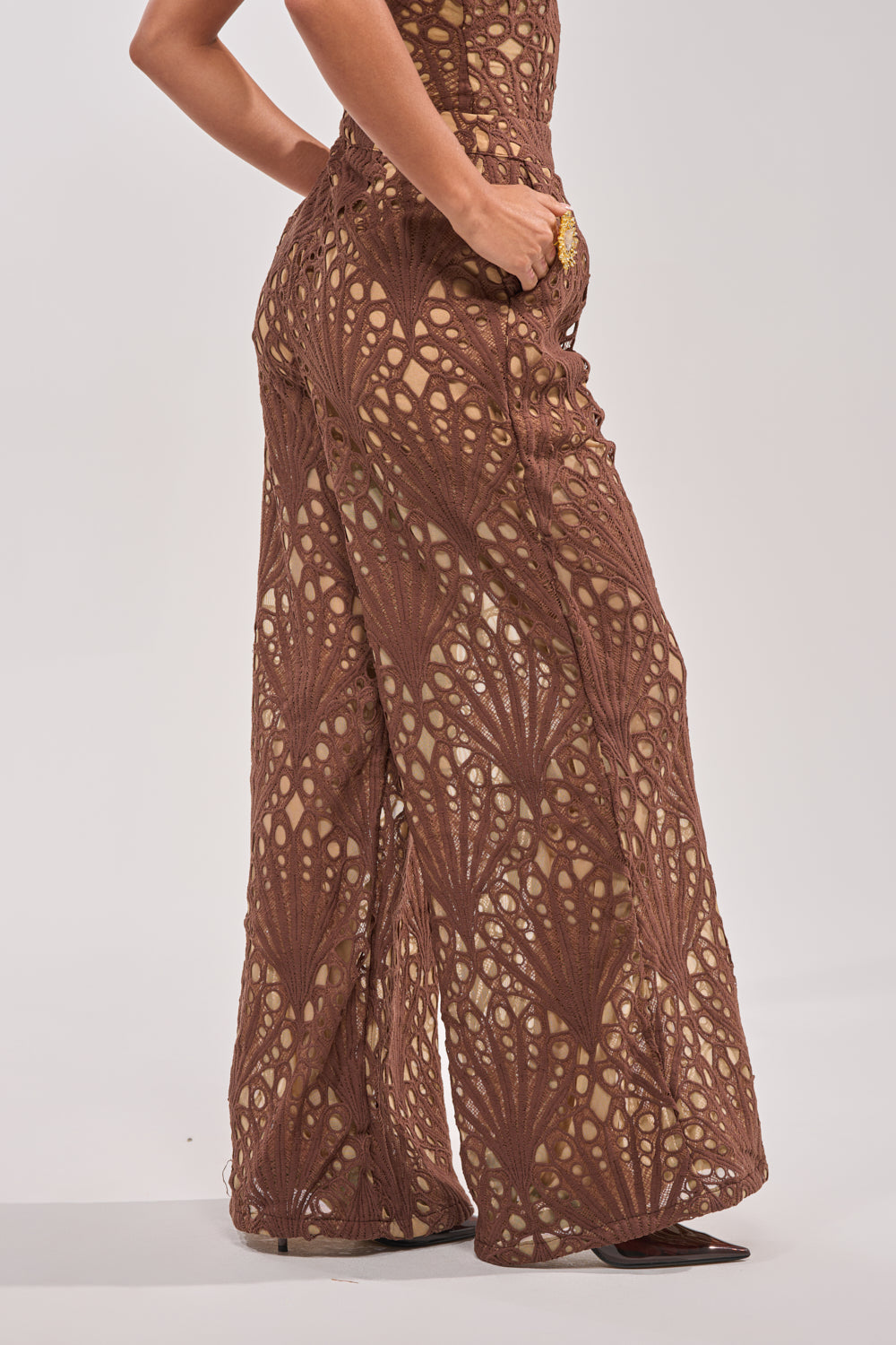 Wearing the CLAIRE EYELET PALAZZO PANT, featuring a fan-like eyelet design over beige lining, a person poses with one hand in their pocket against a plain light background. 3