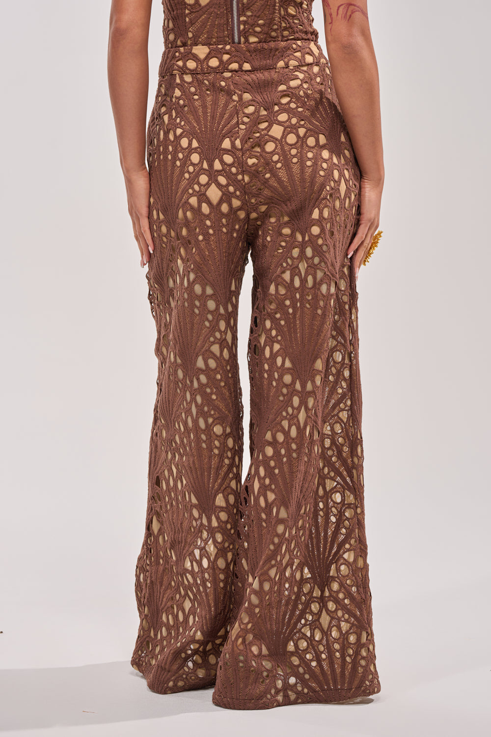 A person is seen from the back wearing the CLAIRE EYELET PALAZZO PANT—brown, wide-leg lace pants with an intricate eyelet cutout pattern over nude lining, set against a light background. 4
